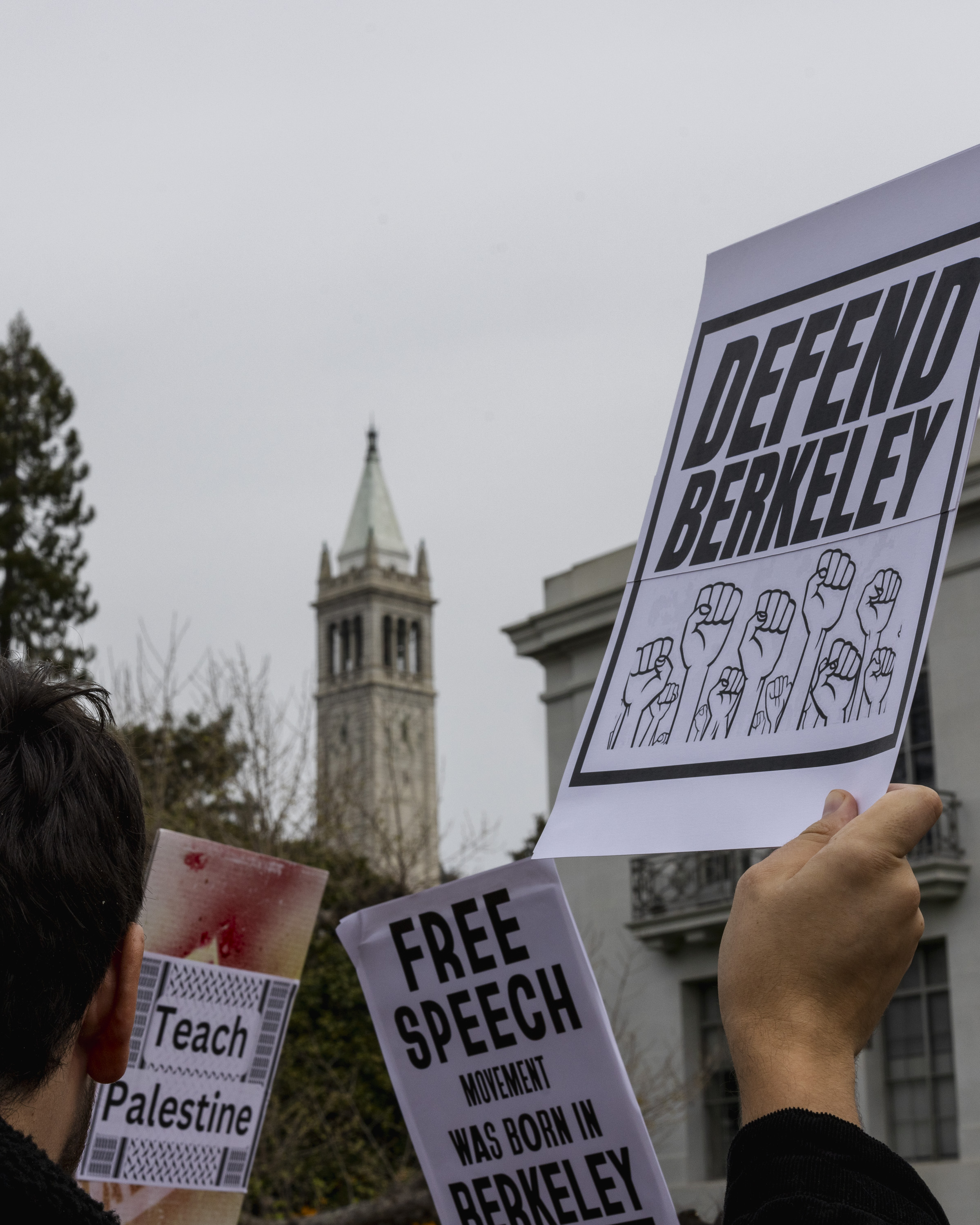 People hold protest signs reading &quot;Defend Berkeley,&quot; &quot;Teach Palestine,&quot; and &quot;Free speech movement was born in Berkeley,&quot; with a tower in the background.