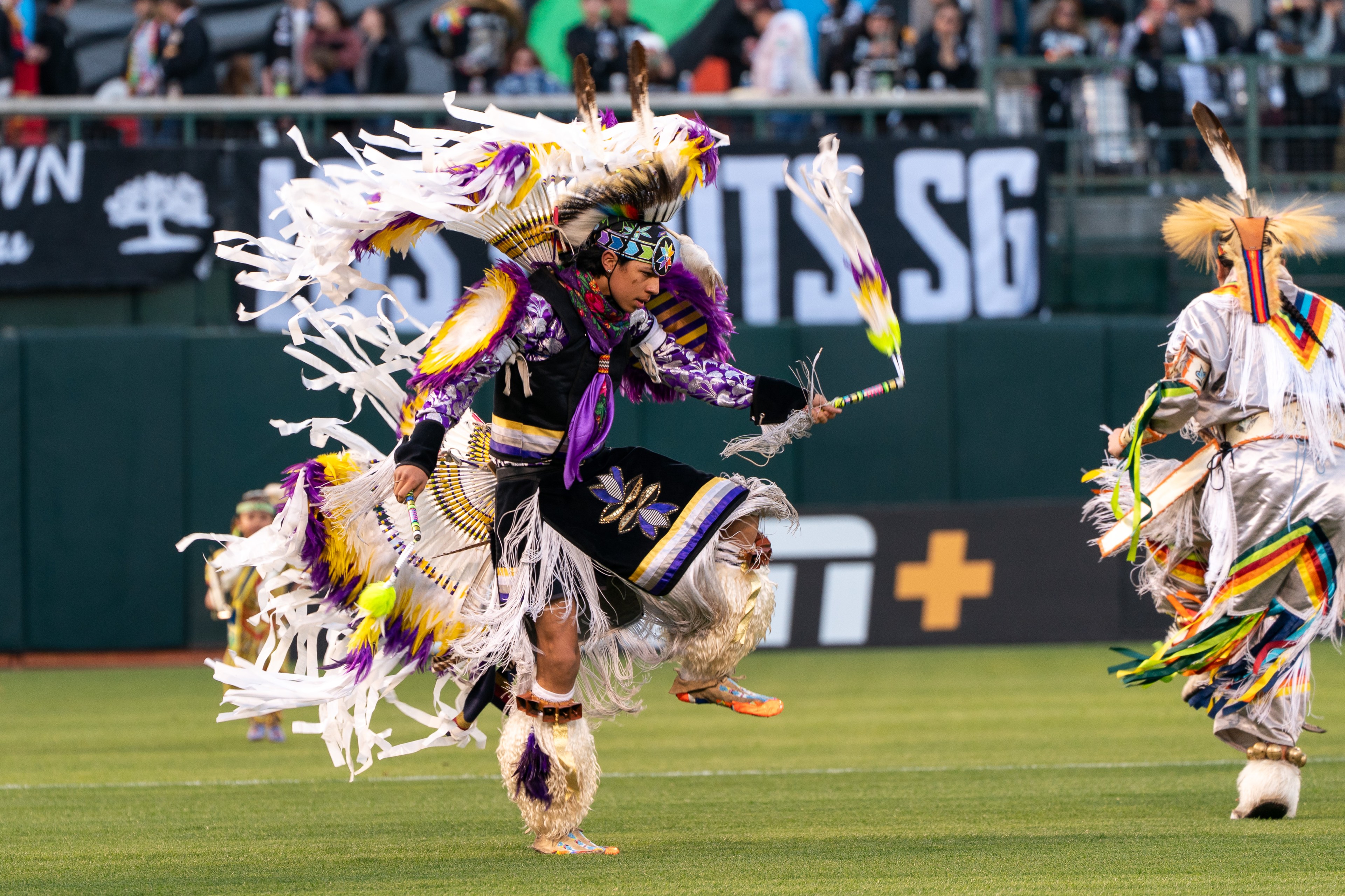 A person in vibrant, feathered regalia dances energetically on a grass field, surrounded by a cheering crowd in the background.