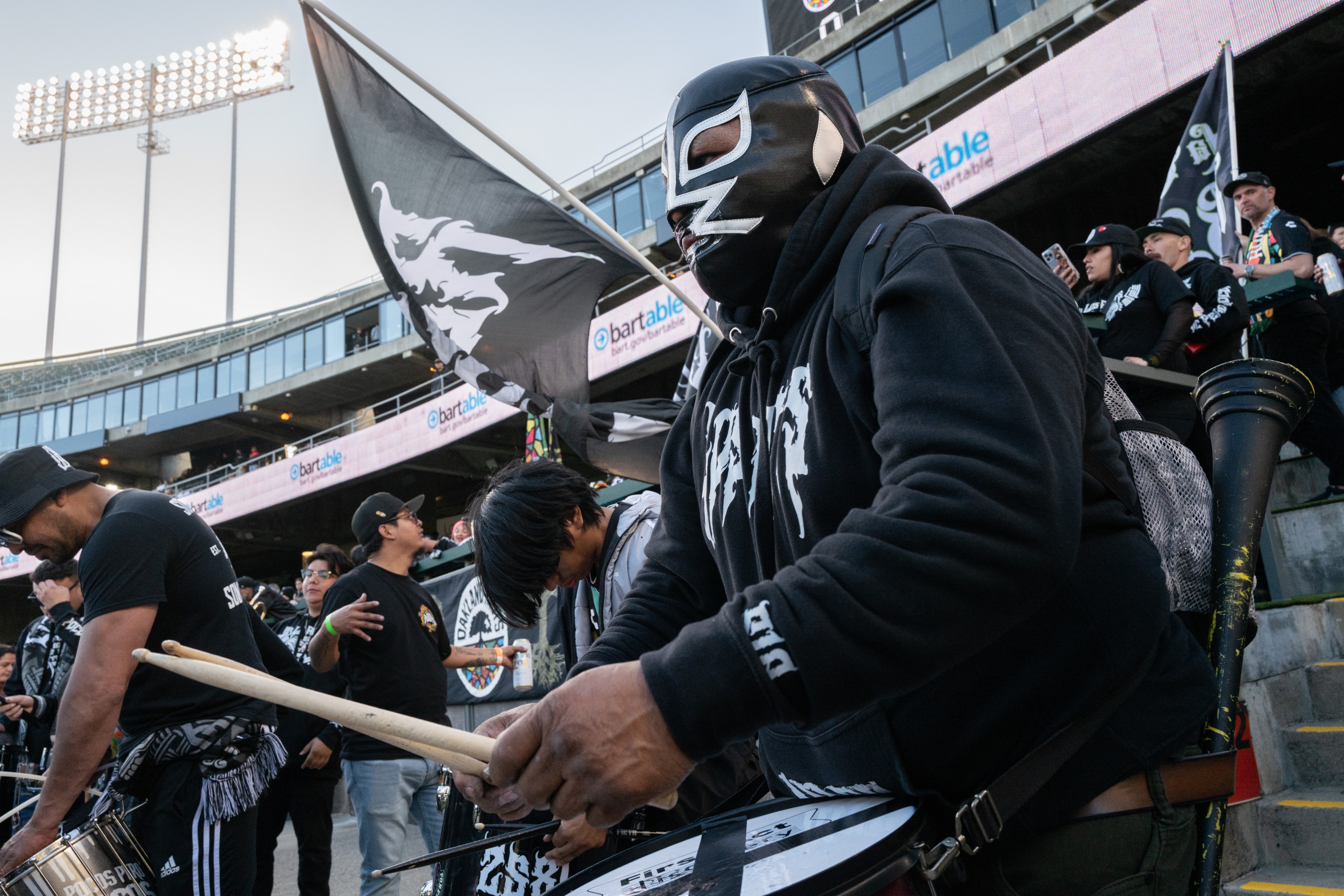 A person in a black mask and hoodie plays a drum in a lively crowd, holding flags. They're in a stadium with lights above, surrounded by cheering fans.