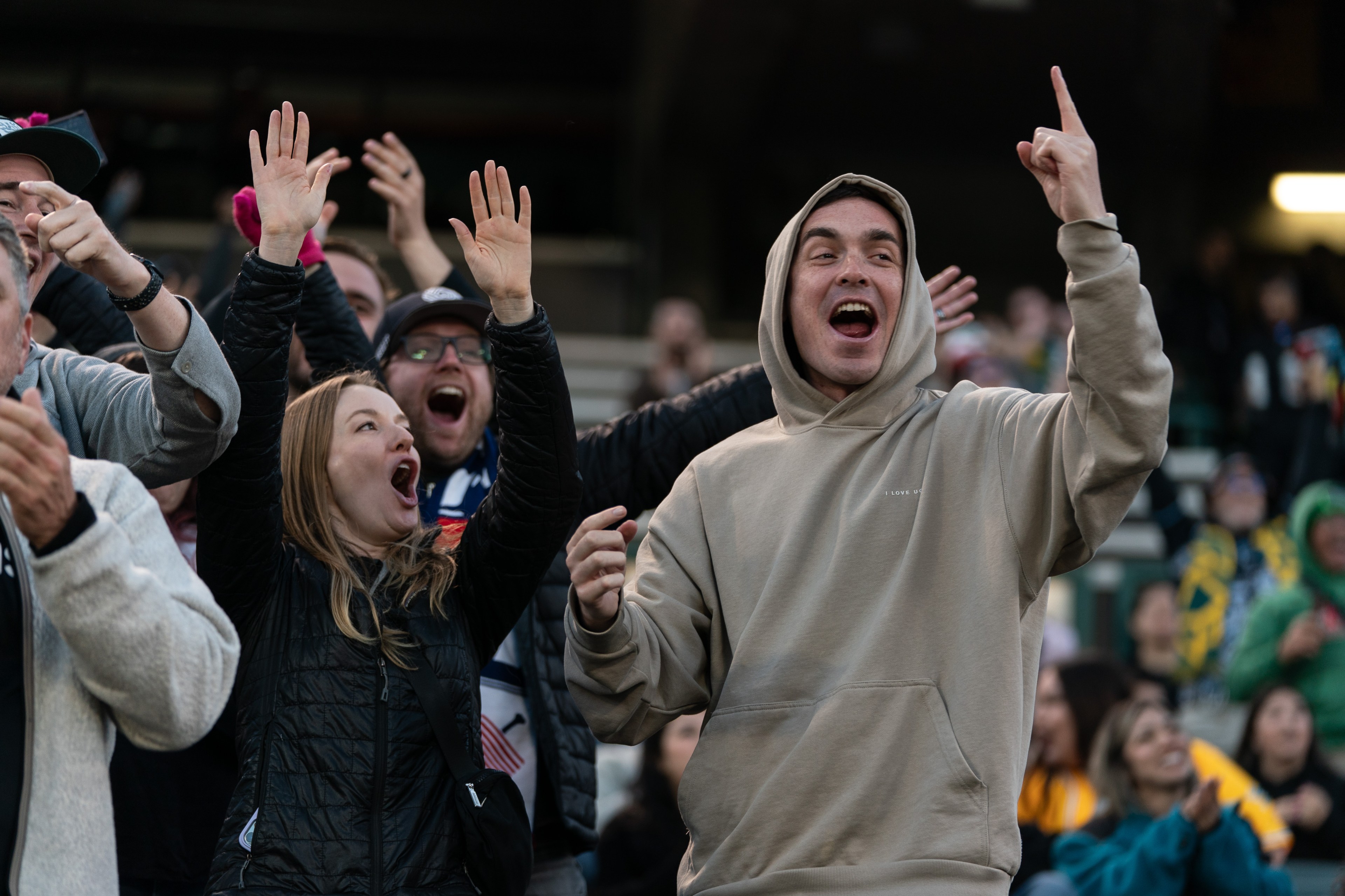 A group of excited fans is cheering energetically at an event. One person in a tan hoodie is pointing upwards, while others are raising their hands and shouting.