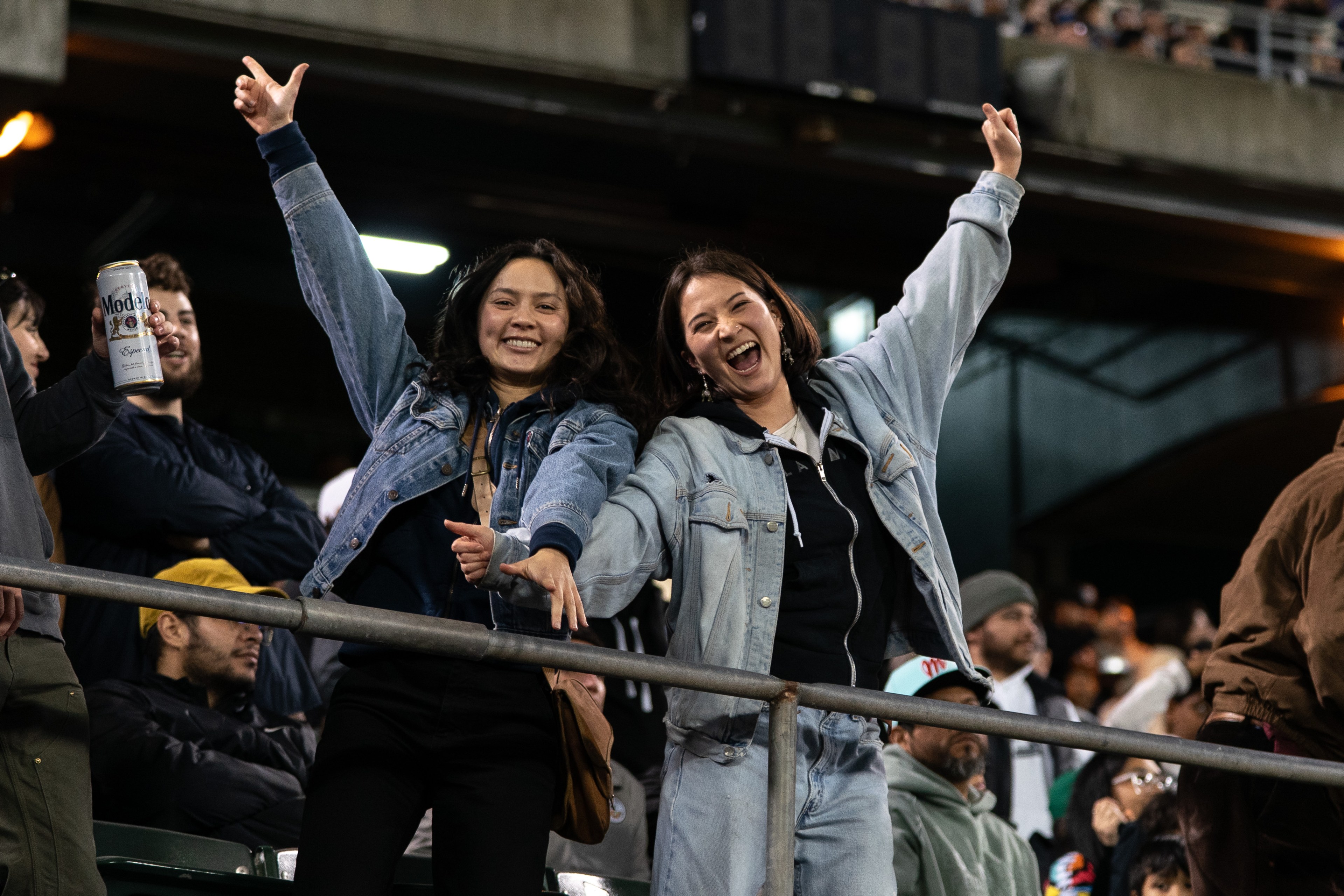 Two people in denim jackets enthusiastically pose with arms raised in a lively crowd. One holds a can, while others smile in the background.