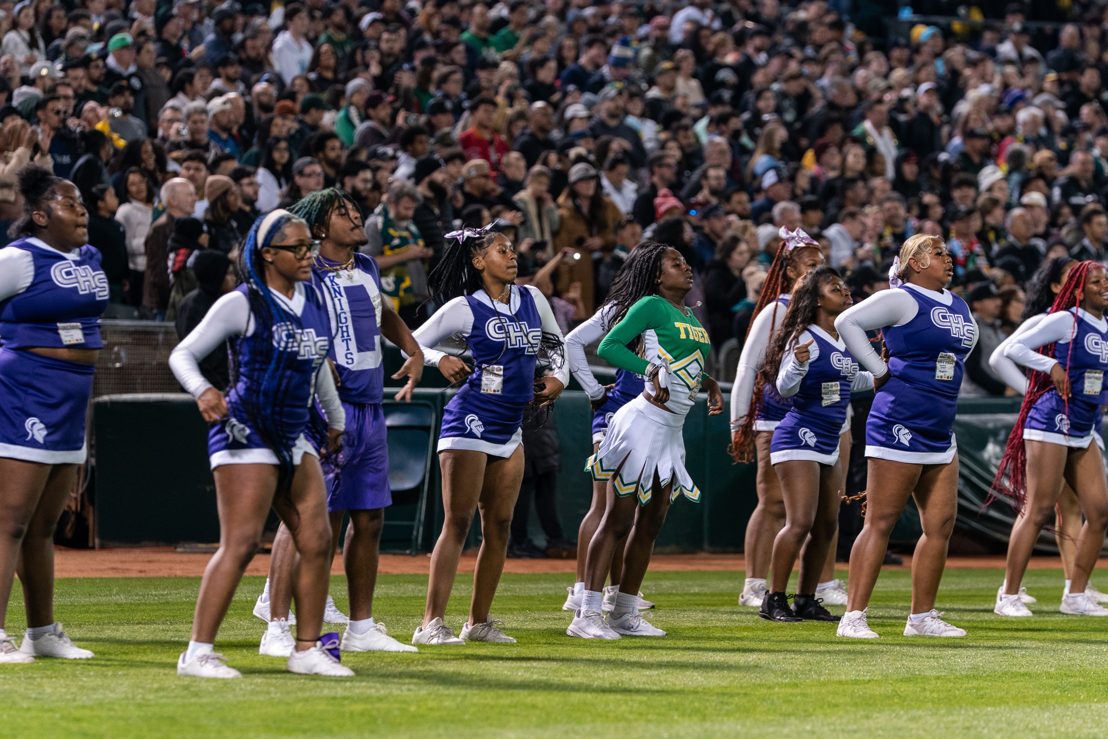 A group of cheerleaders is performing on a field, wearing blue and purple uniforms marked &quot;CHS,&quot; and one in green and yellow. A crowd watches in the background.