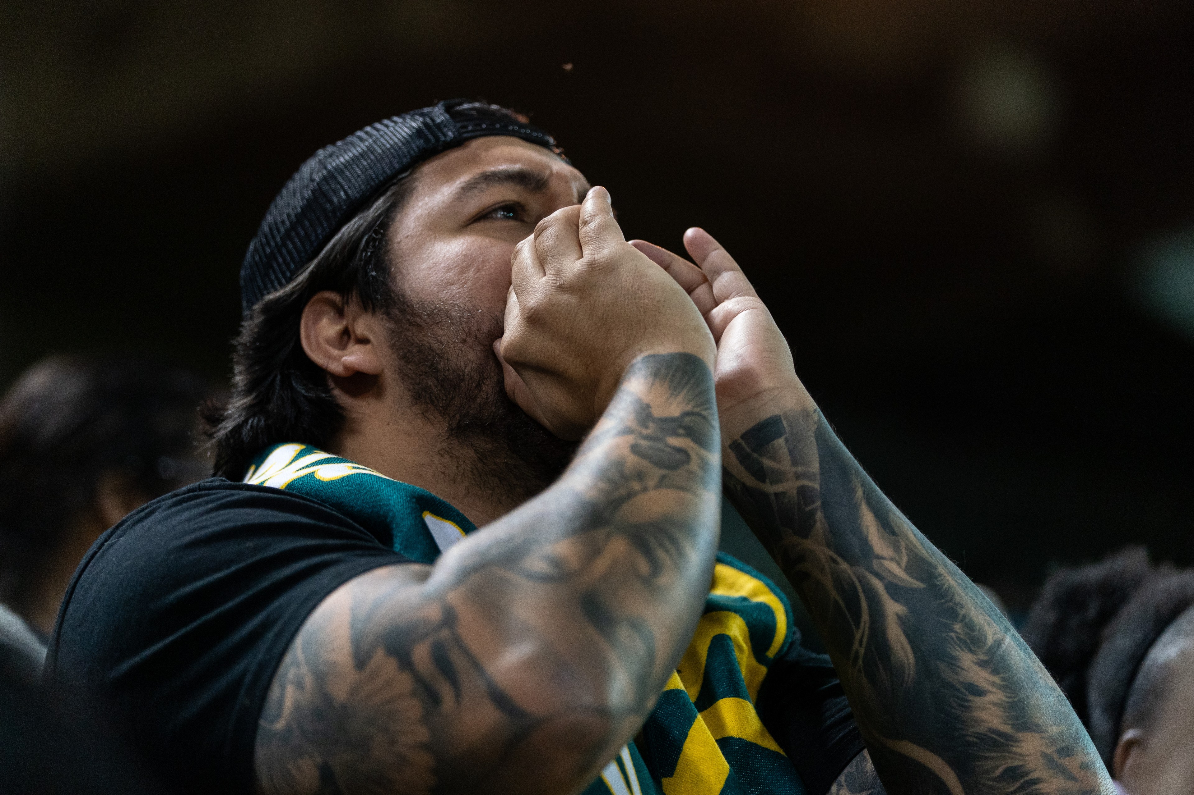 A person with tattoos on their arms and wearing a backward cap holds their hands to their mouth as if shouting, with a green and yellow scarf around their neck.