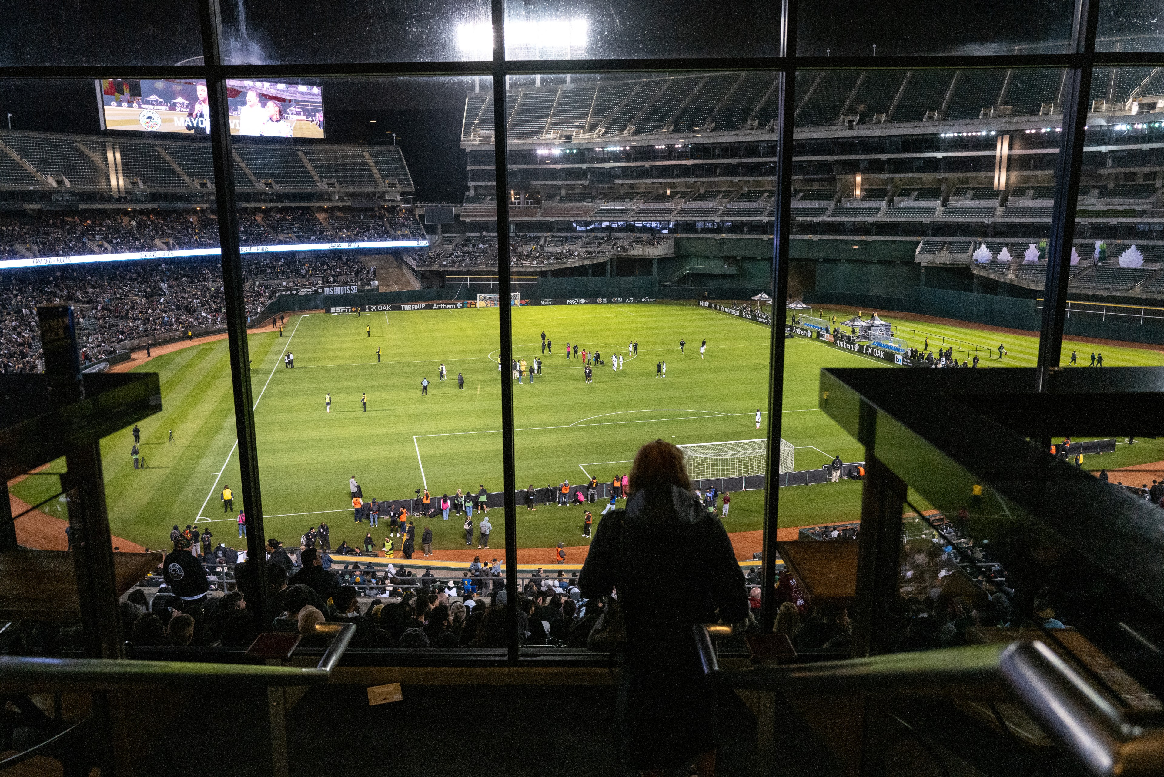 A stadium filled with spectators overlooks a soccer field. People are positioned around the field, and a large screen displays visuals to the audience.