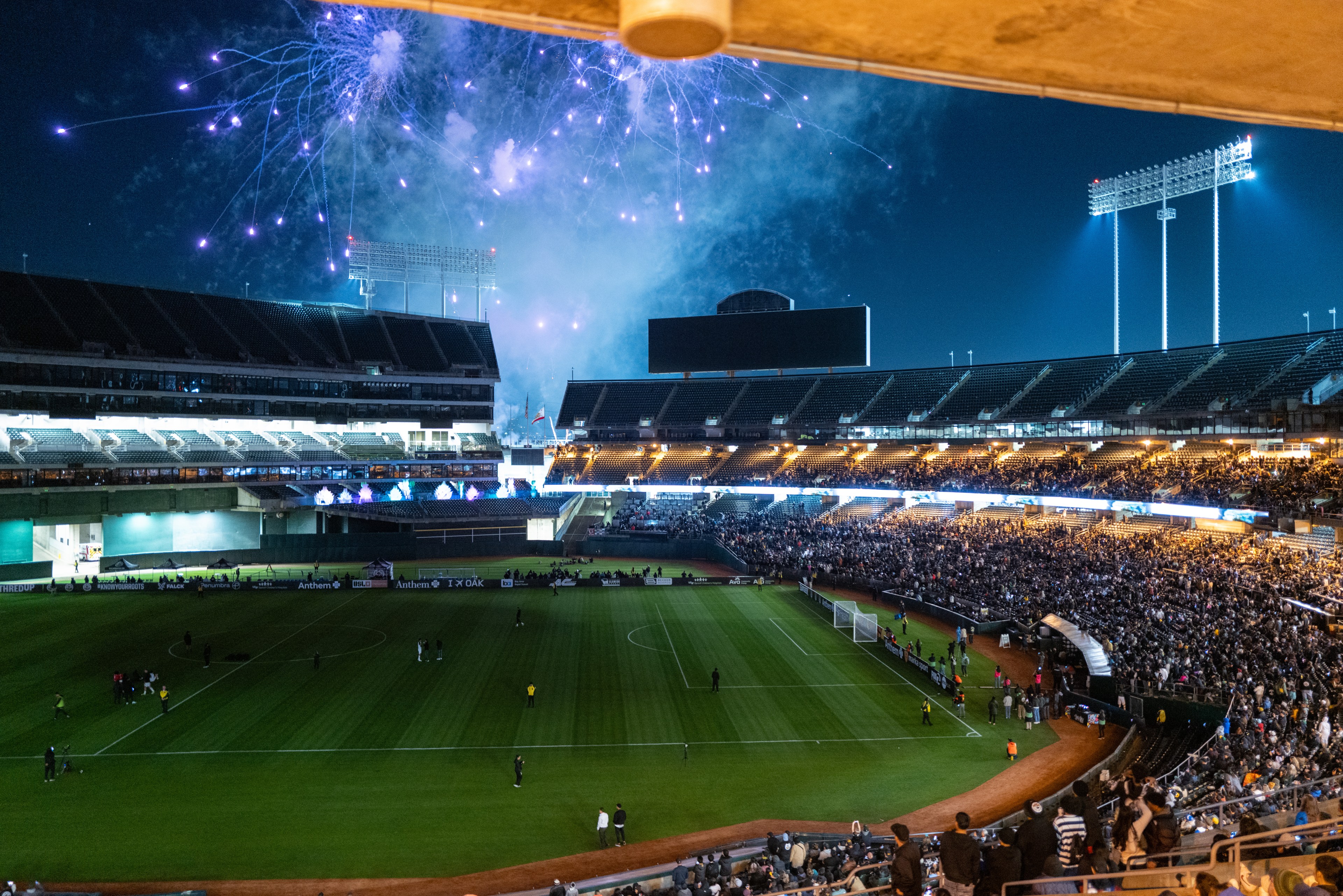 A stadium lit up with fireworks in the night sky, with a crowd of spectators around a soccer field, creating a festive atmosphere.