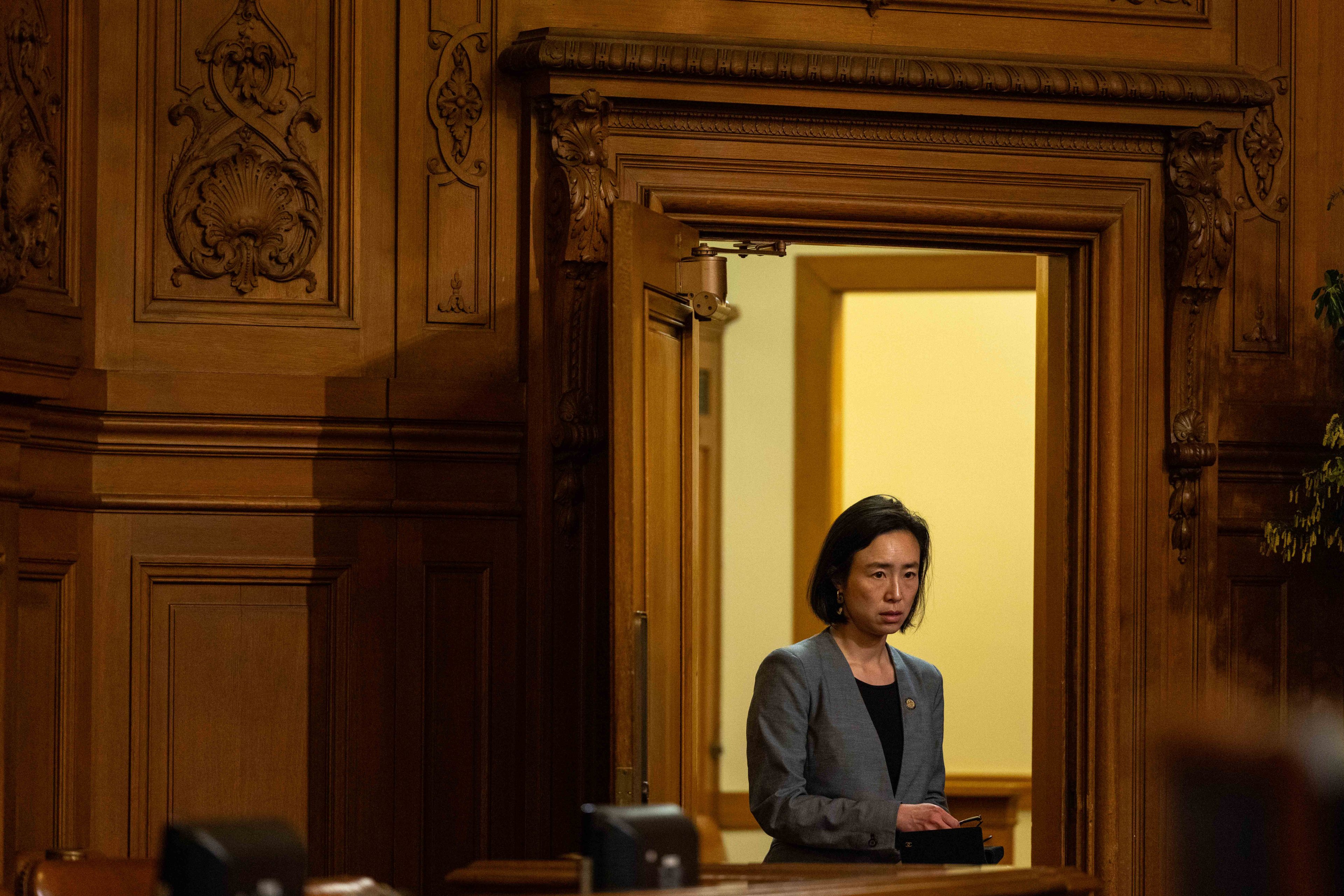 A person in a gray jacket is standing in a richly carved wooden doorway, holding a black item, looking forward with a neutral expression.