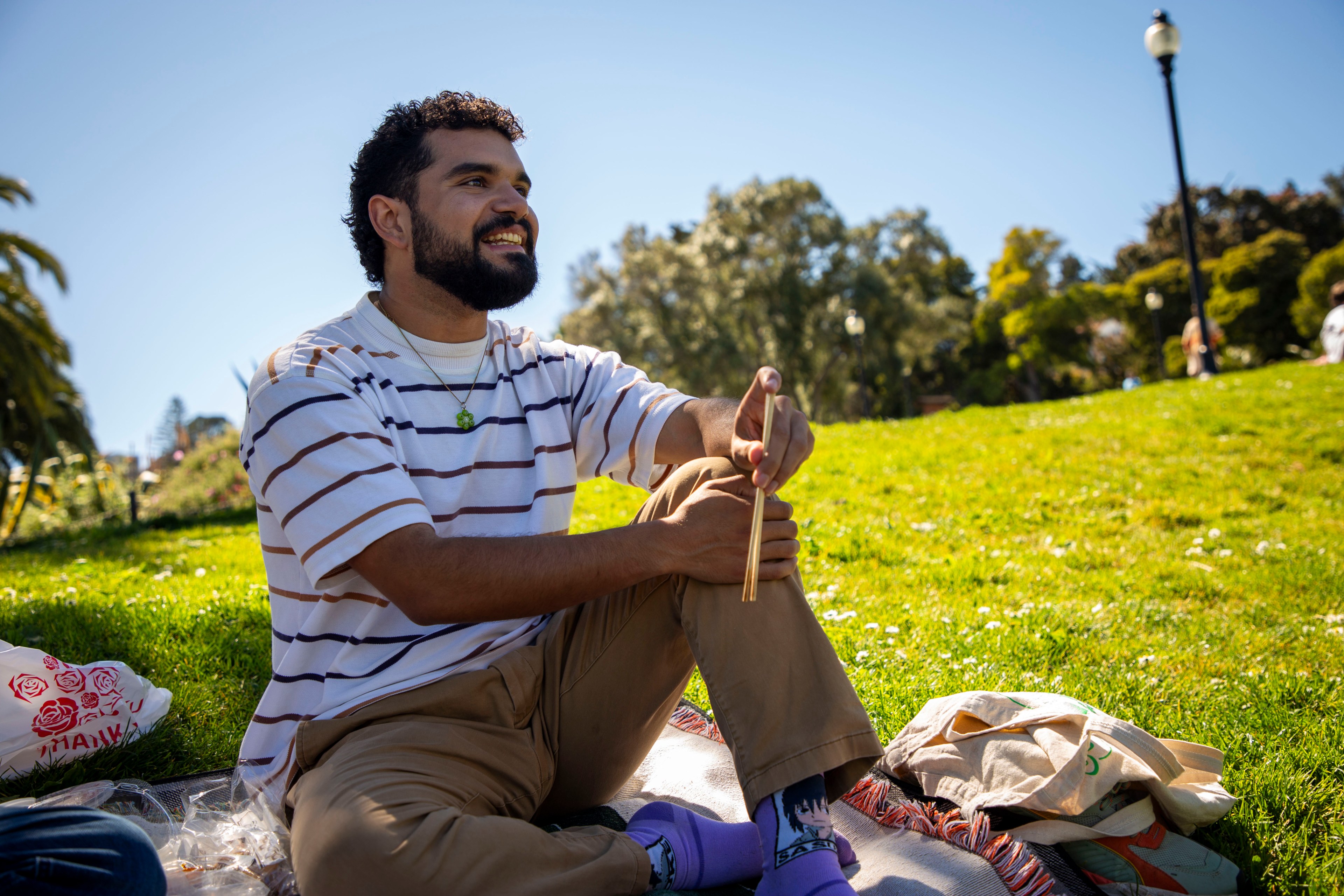 A person sits on a blanket in a sunny park, smiling and holding chopsticks. They wear a striped shirt, brown pants, and purple socks. A lamp post is in the background.