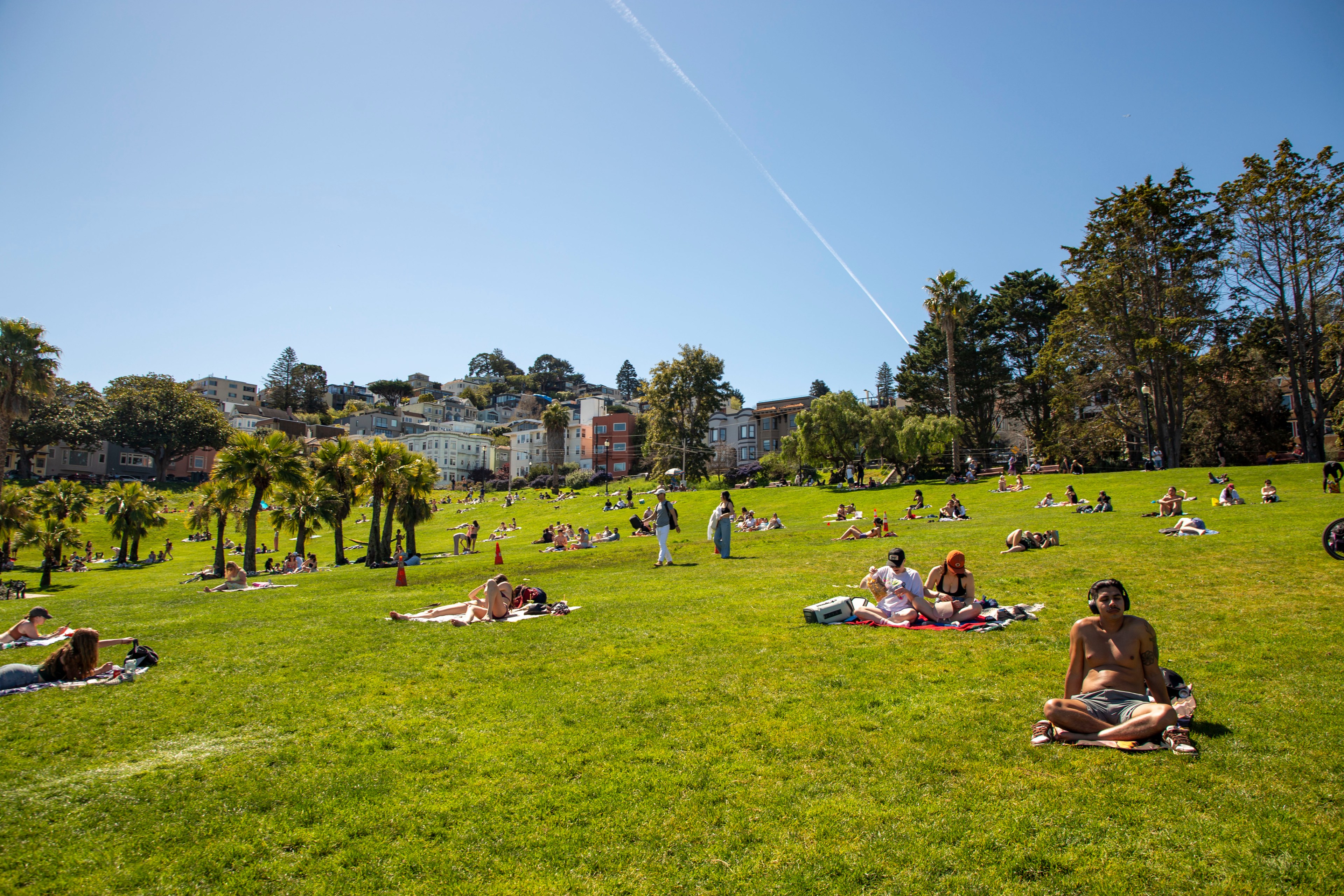 A sunny park scene shows people relaxing on a grassy hill with clear blue skies. Some are on blankets, and colorful houses are visible in the background.