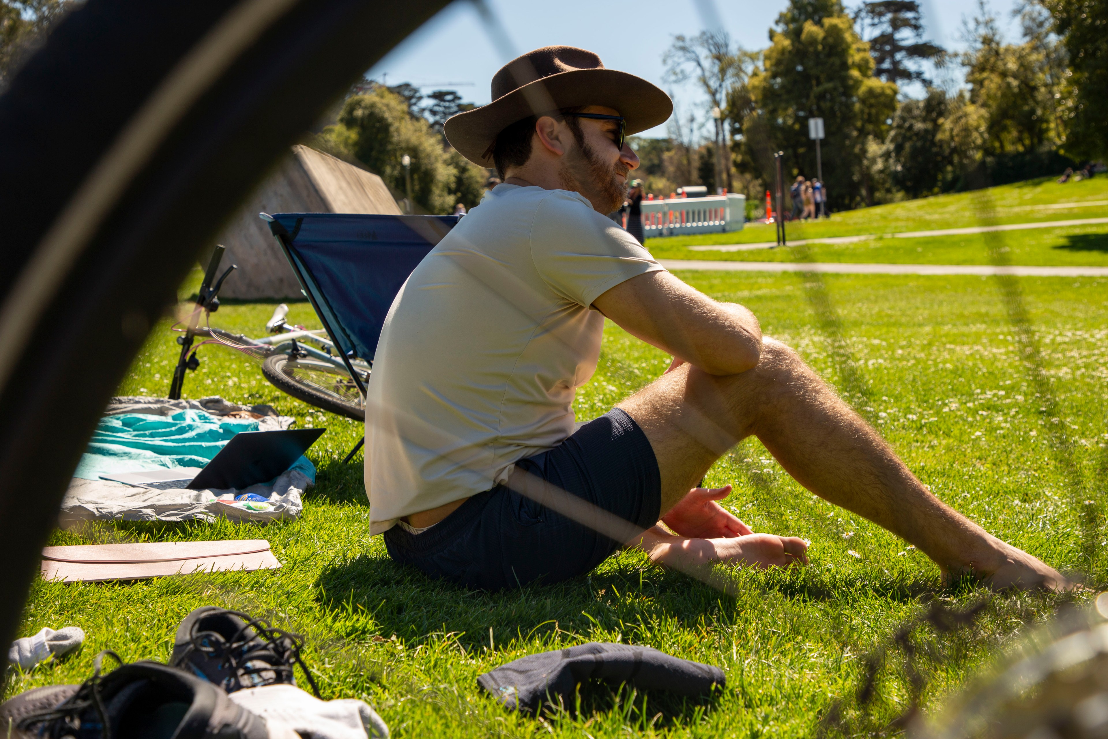 A person wearing a hat and sunglasses sits on grass in a park, surrounded by a bike, shoes, and a blue chair under sunny skies.