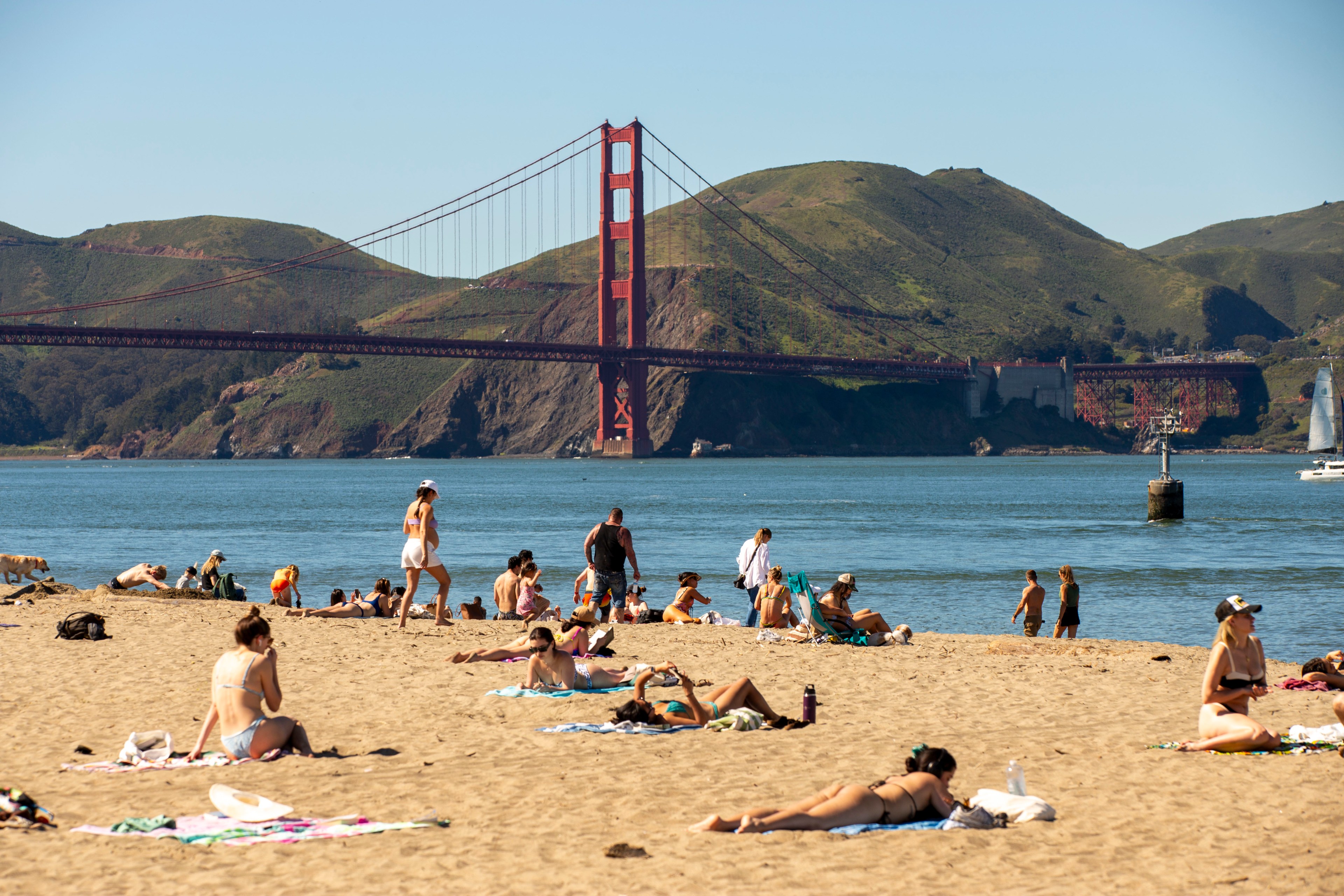 People relax on a sandy beach with the iconic Golden Gate Bridge and green hills in the background. A sunny day with calm water and a few sailboats.