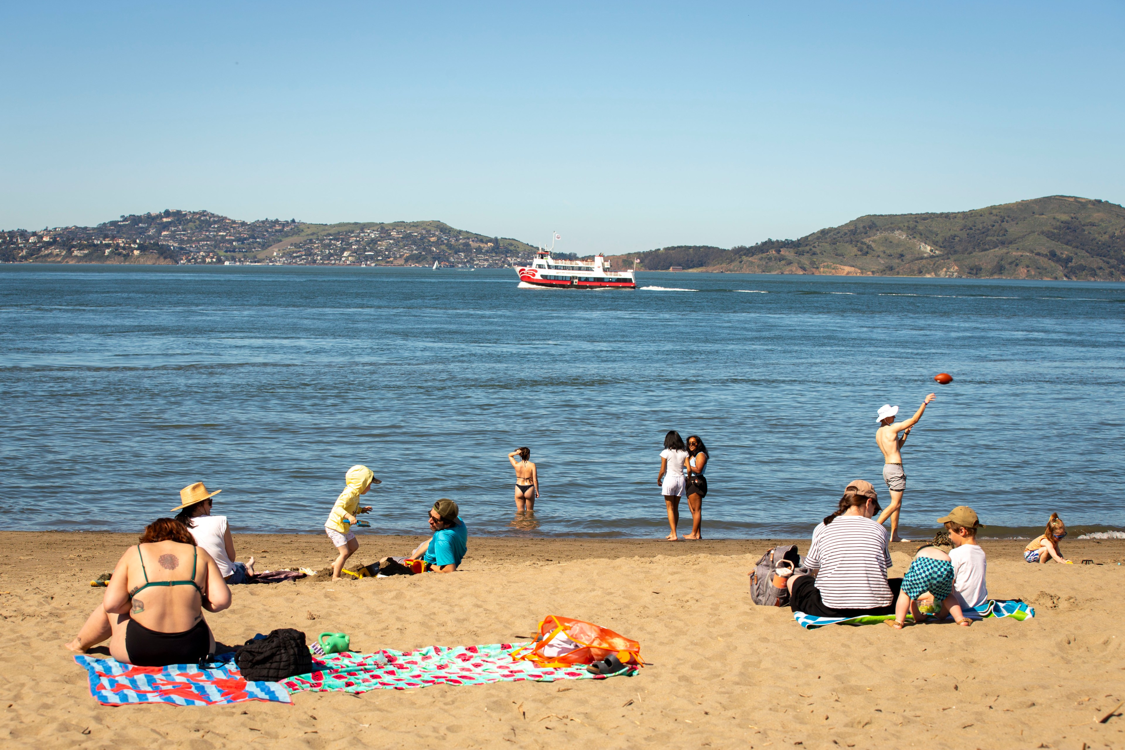 People relax on a sandy beach with towels under clear skies. Some are near the water, while a red and white boat passes by on the sea, with hills in the background.