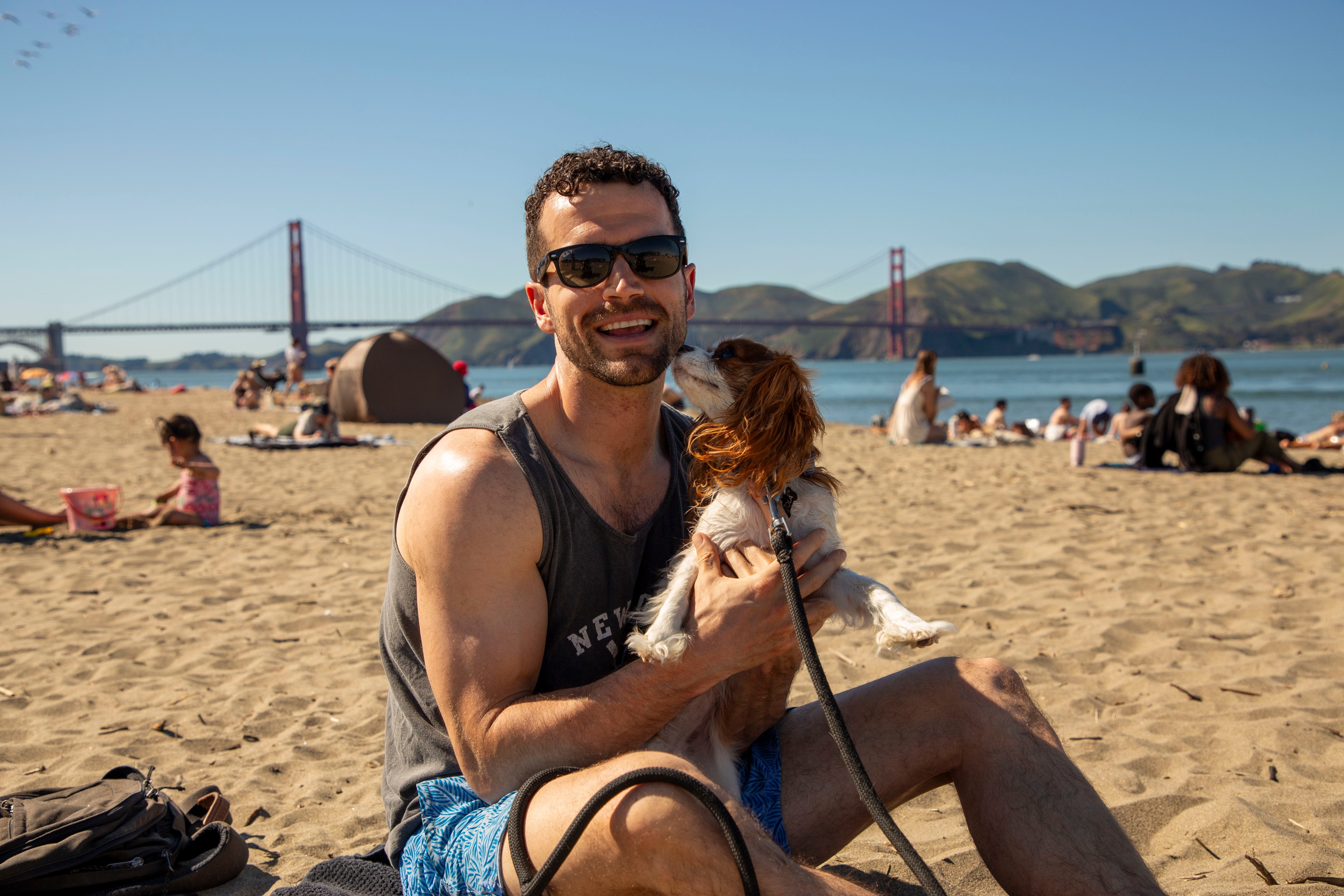 A smiling man in sunglasses is sitting on a sunny beach holding a small dog. The Golden Gate Bridge is visible in the background with people lounging around.