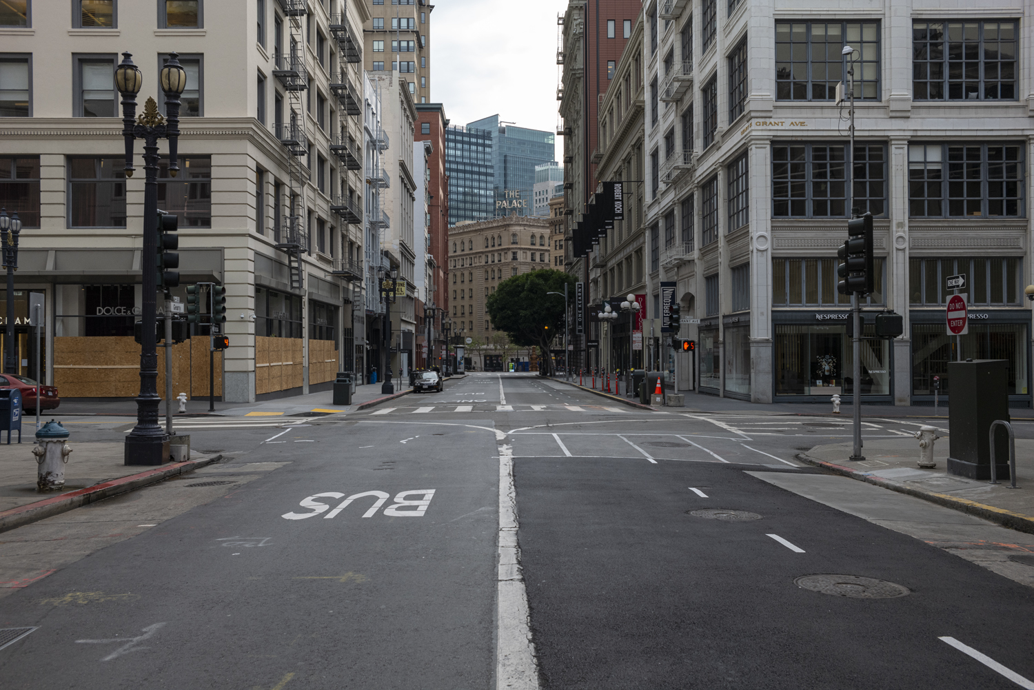 An empty city street with tall buildings on both sides, boarded shops, and traffic signals. &quot;The Palace&quot; sign is visible in the background.