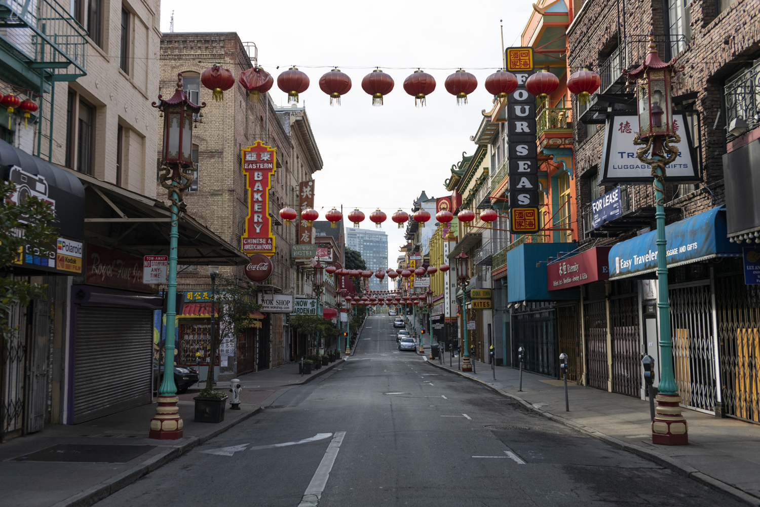 A narrow street lined with shops in a Chinatown setting, featuring red lanterns hanging overhead, colorful signs in English and Chinese, and some parked cars.