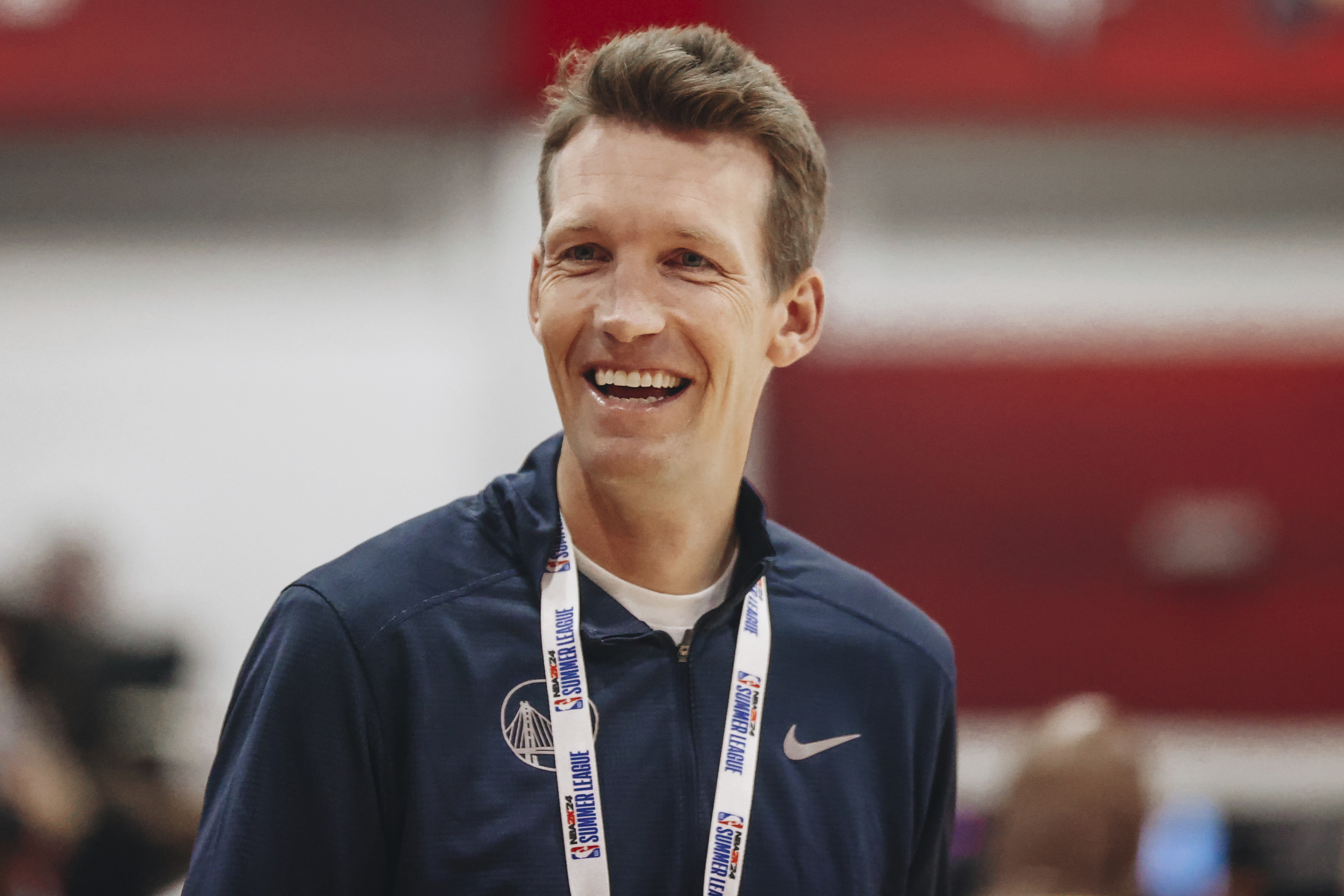 A person is smiling broadly while wearing a dark jacket with a lanyard around their neck. The background is blurred, showing a red wall and indistinct shapes.