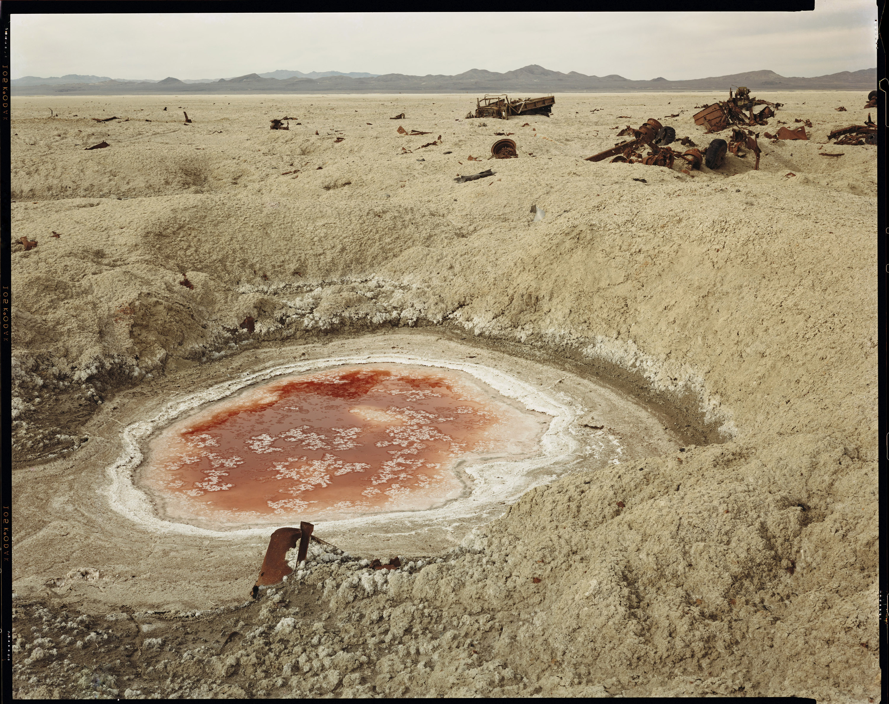 A desolate landscape with a red, circular pond surrounded by crusty salt flats and scattered rusty debris, set against distant mountains.