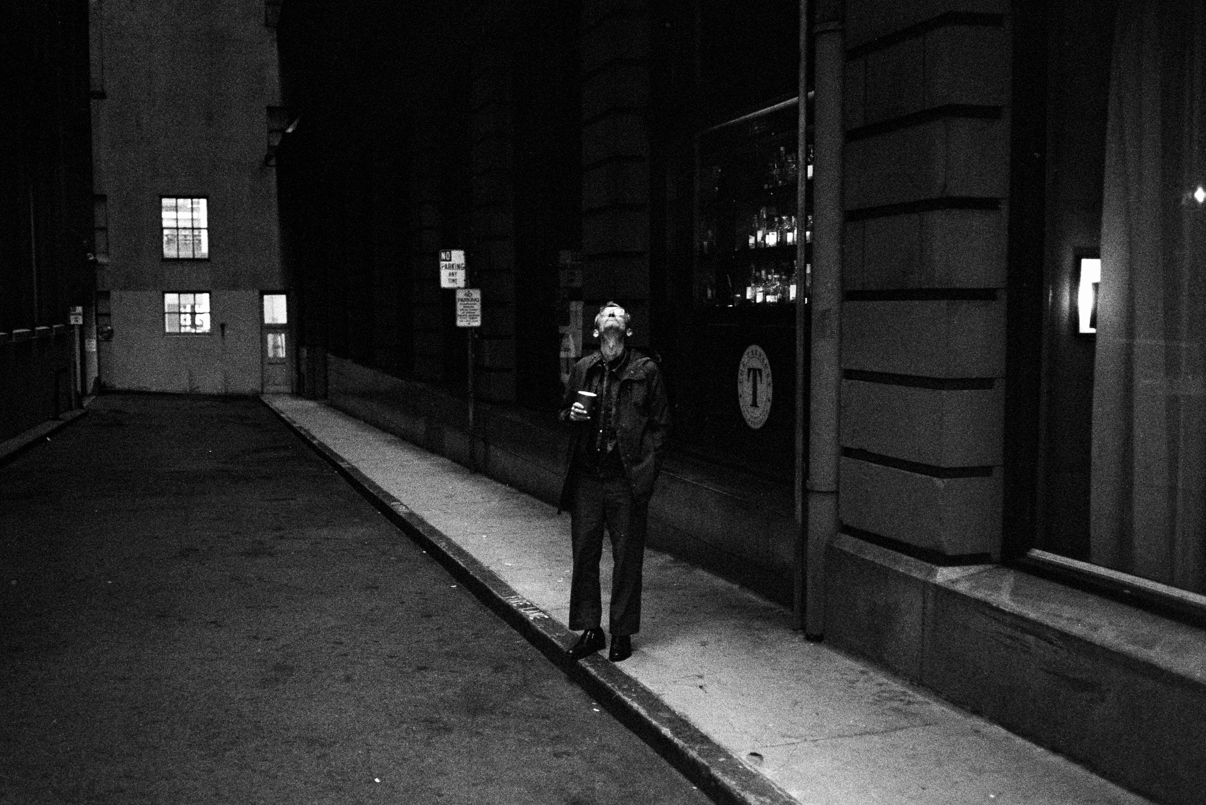 A person stands on a dimly lit sidewalk next to a building at night, gazing upward while holding a drink. There's a &quot;No Parking&quot; sign nearby.