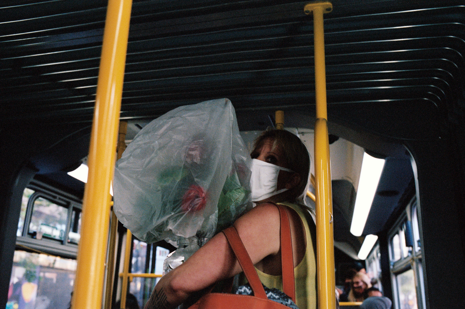 A woman on a bus is holding a bouquet covered in plastic. She wears a mask and a sleeveless top, with a brown bag on her shoulder. The bus interior is visible.