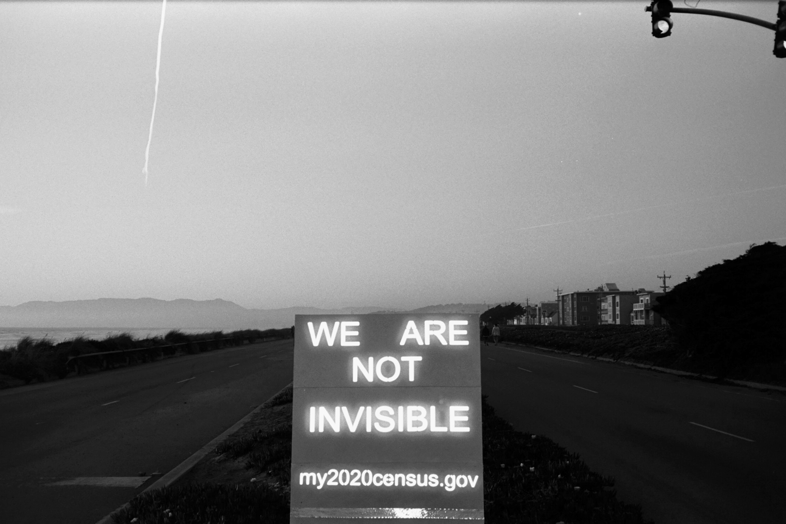 A sign on an empty roadside reads &quot;WE ARE NOT INVISIBLE&quot; with &quot;my2020census.gov&quot; below. It's evening; buildings and distant hills are visible.