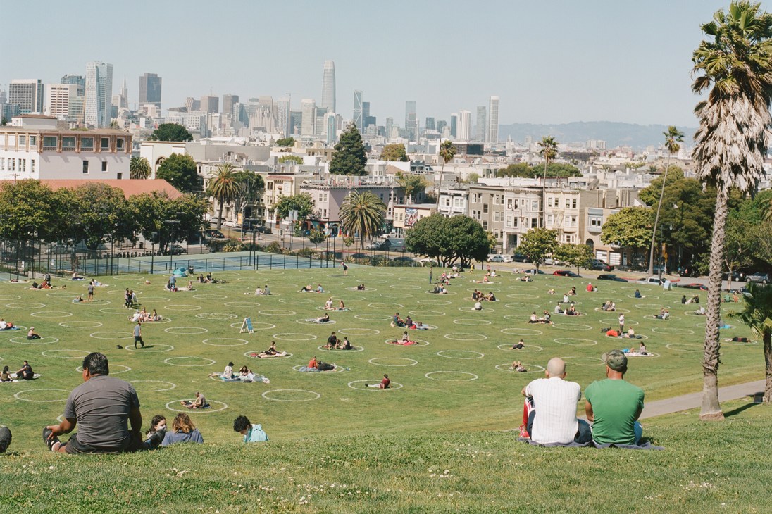 People are enjoying a sunny day in a spacious park, sitting in distanced circles on the grass. The city skyline is visible in the background.