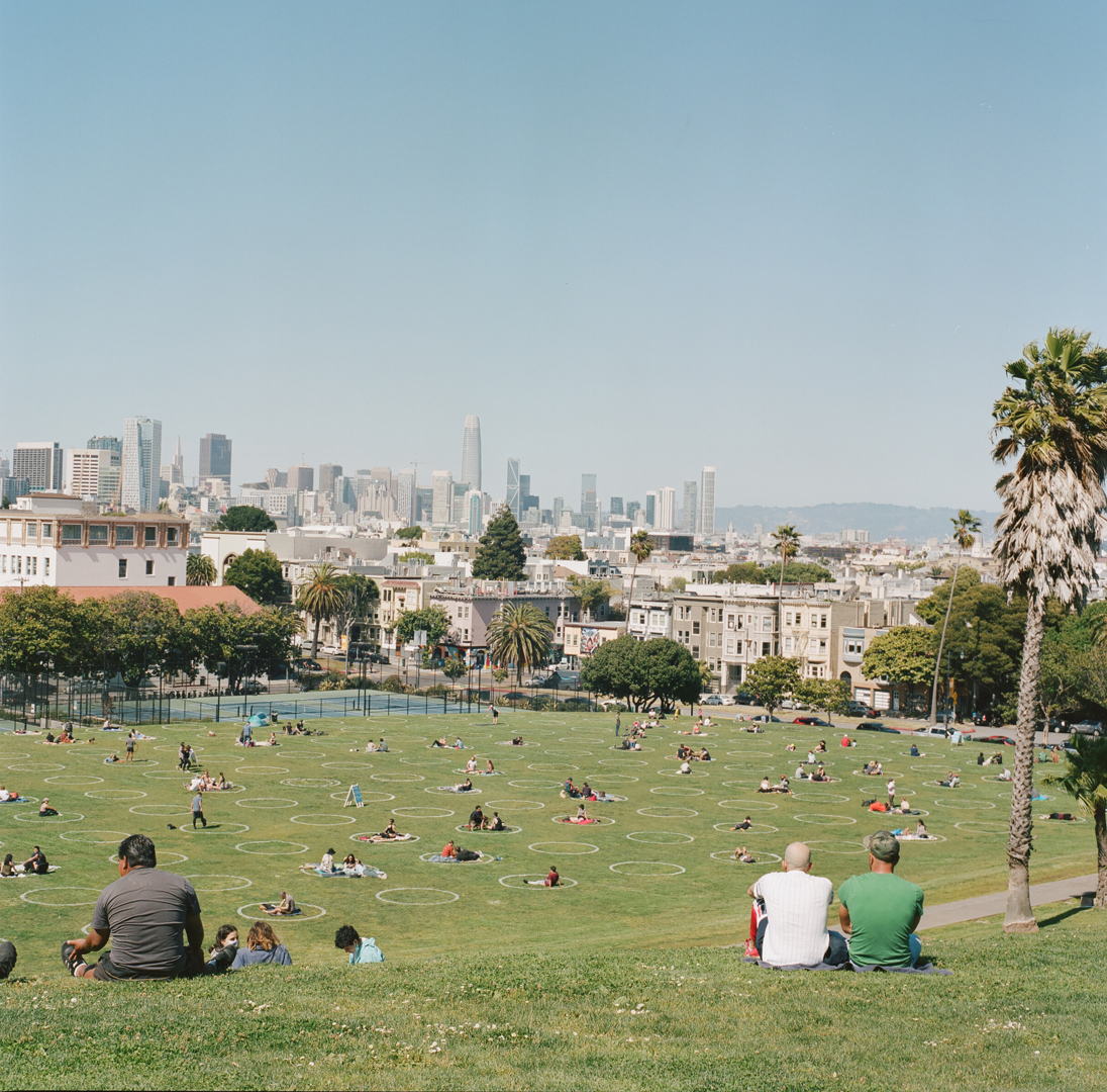 People are enjoying a sunny day in a spacious park, sitting in distanced circles on the grass. The city skyline is visible in the background.