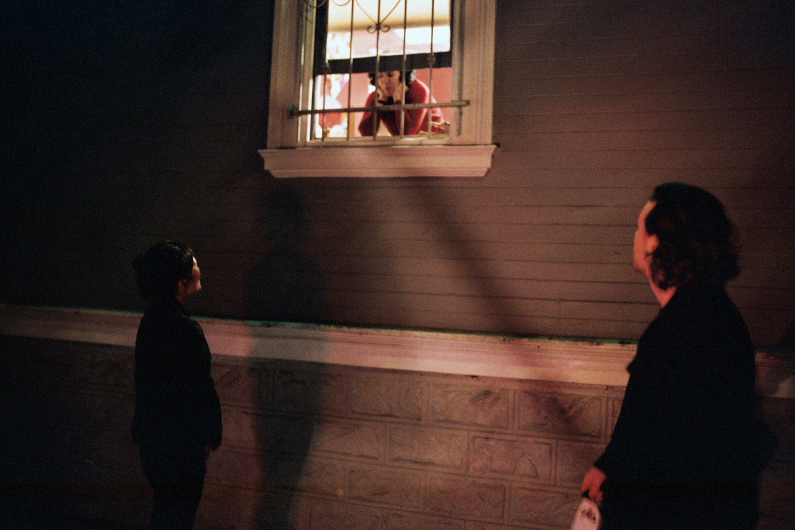 Two people stand outside in the dark, looking up at a person leaning out of a barred window illuminated by warm indoor light.