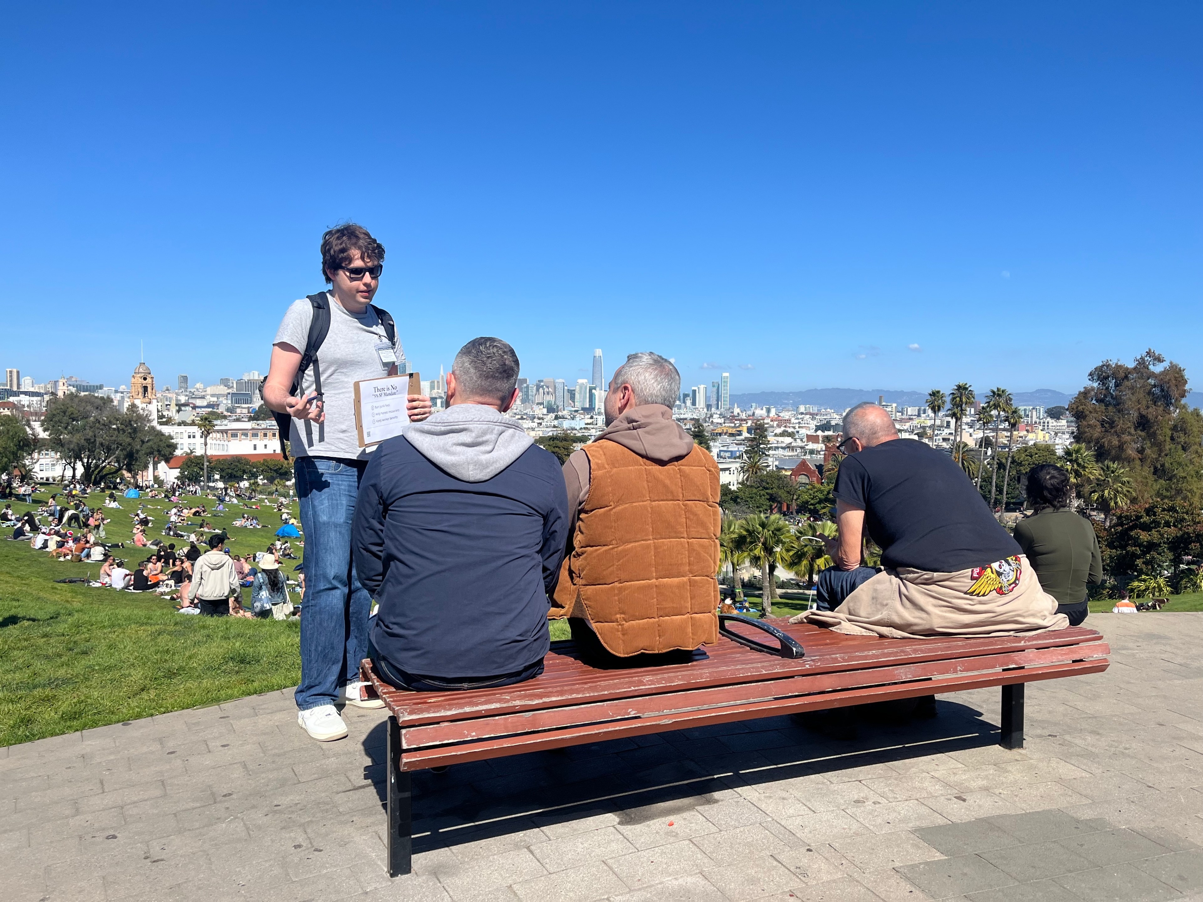 A person stands talking to three seated individuals on a bench, with a park full of people and a city skyline in the background under a clear blue sky.