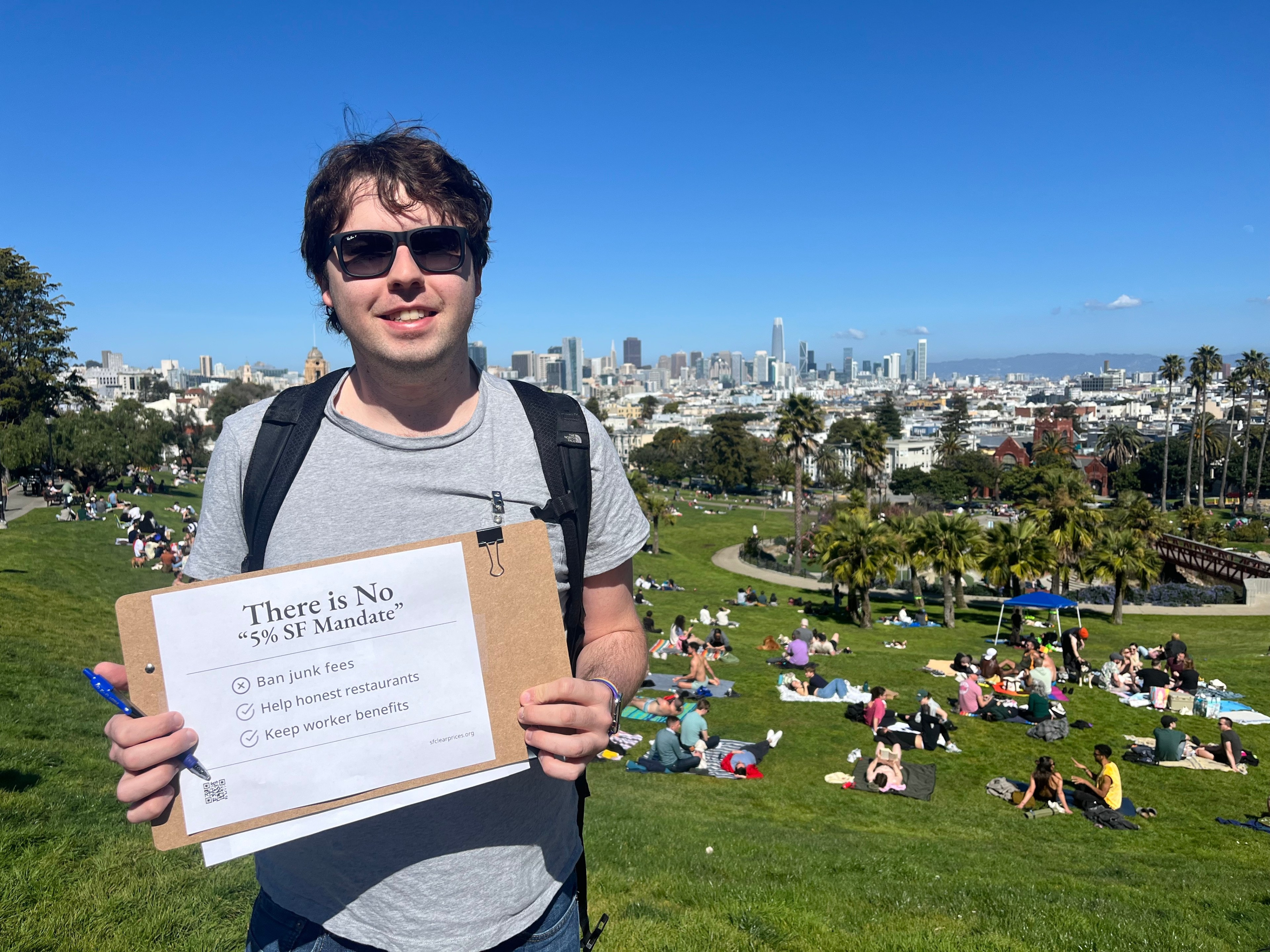 A person wearing sunglasses holds a sign stating "There is No '5% SF Mandate'" in a sunny park. The background shows people relaxing on grass and a city skyline.