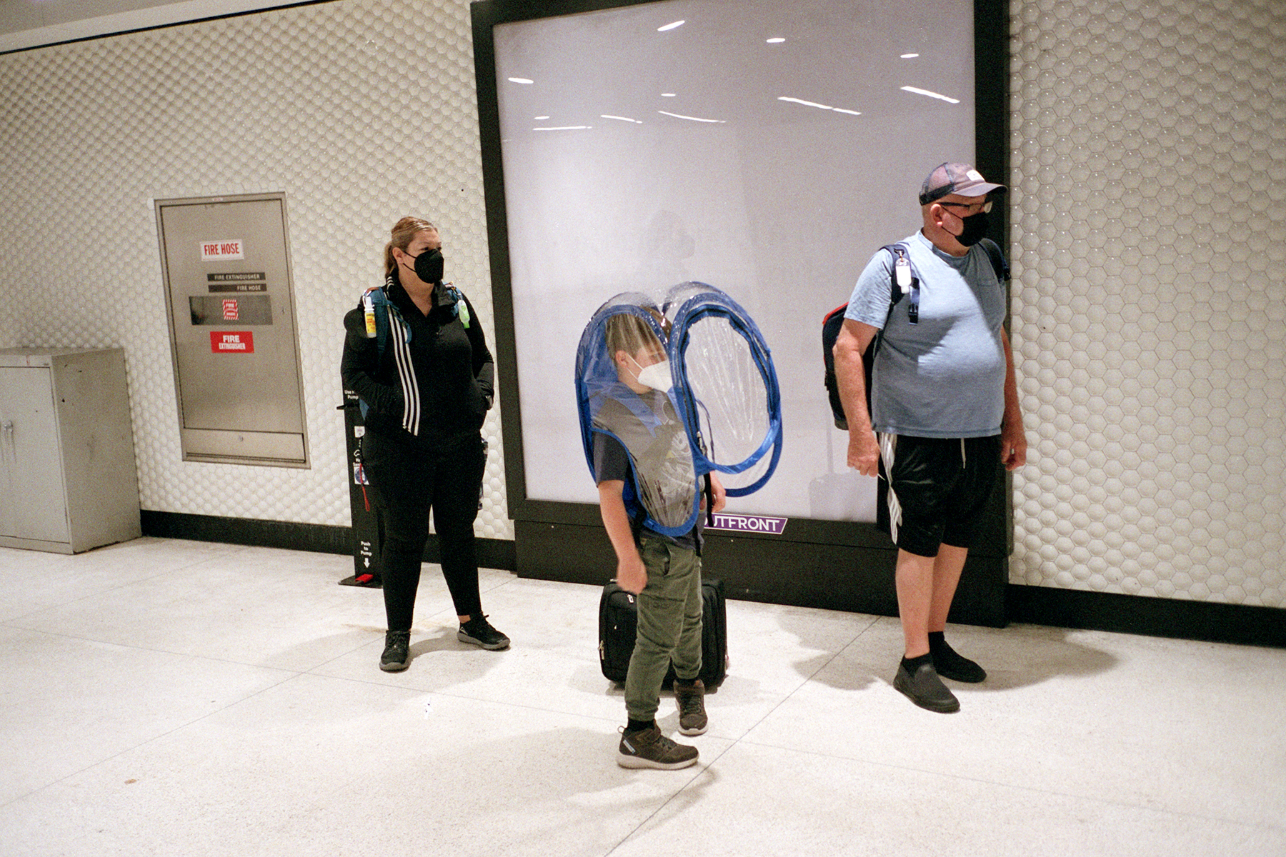 Three people, wearing masks, stand in an indoor area. A child in the center has a transparent protective cover. A fire hose cabinet is visible on the wall.
