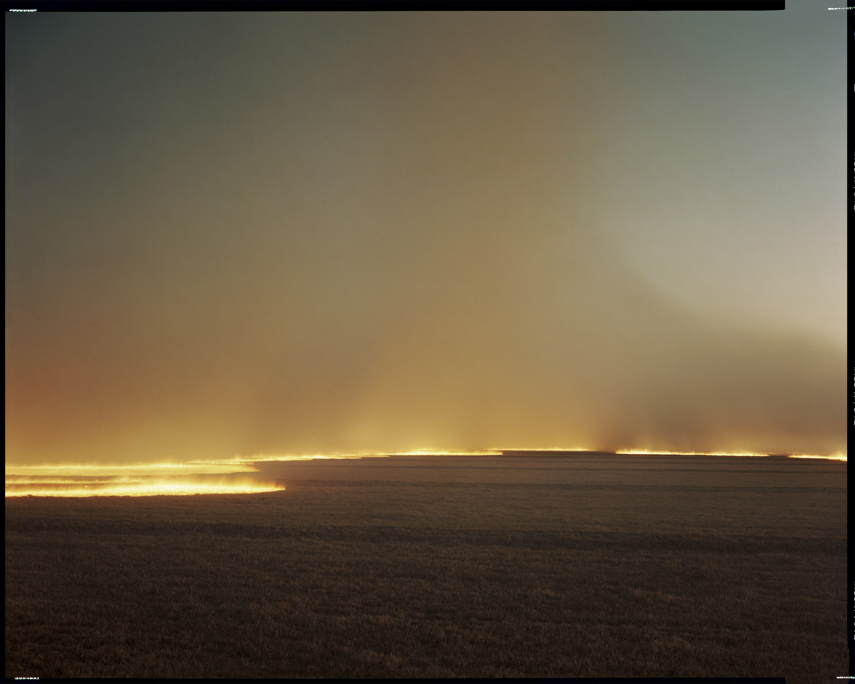 The image shows a vast, flat landscape with a bright horizon of flames, creating a gradient of orange and yellow against a dusky sky.