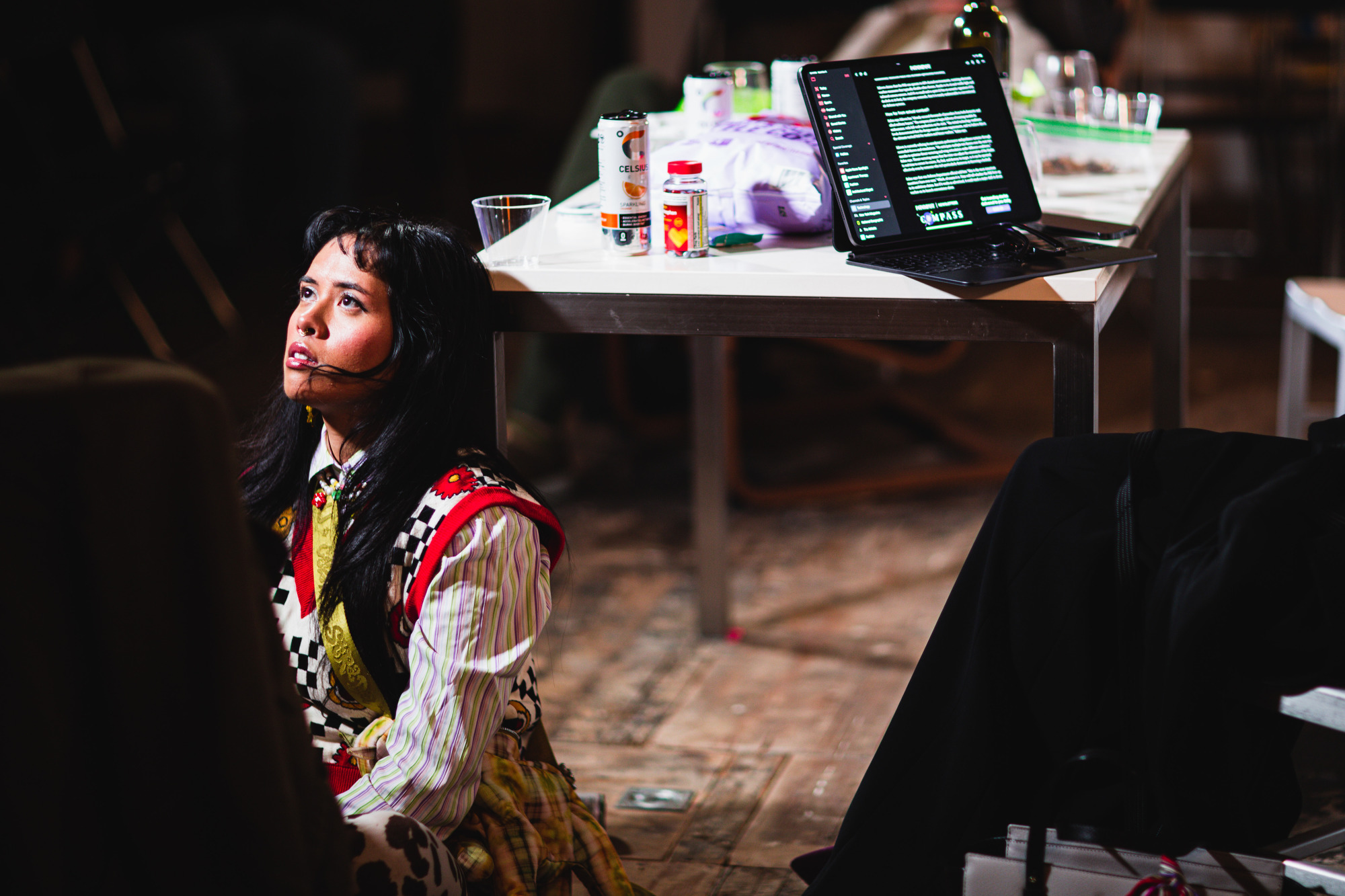 A woman in colorful clothing sits on the floor, gazing upwards. Nearby, a table holds a laptop displaying text, along with drinks and other items.