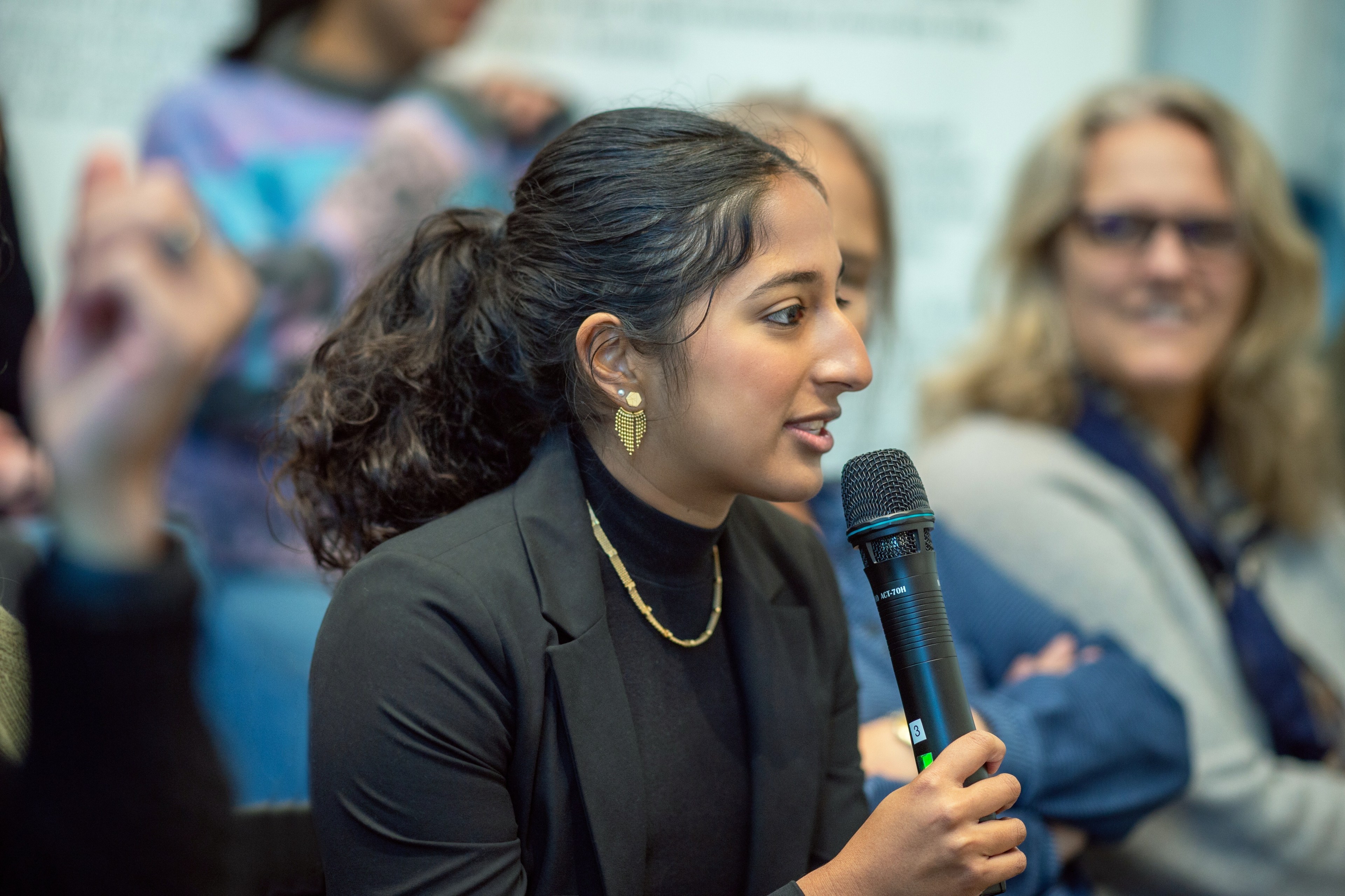 A person with gold earrings and a necklace speaks into a microphone in a group setting. They wear a black outfit, and others are visible in the background.