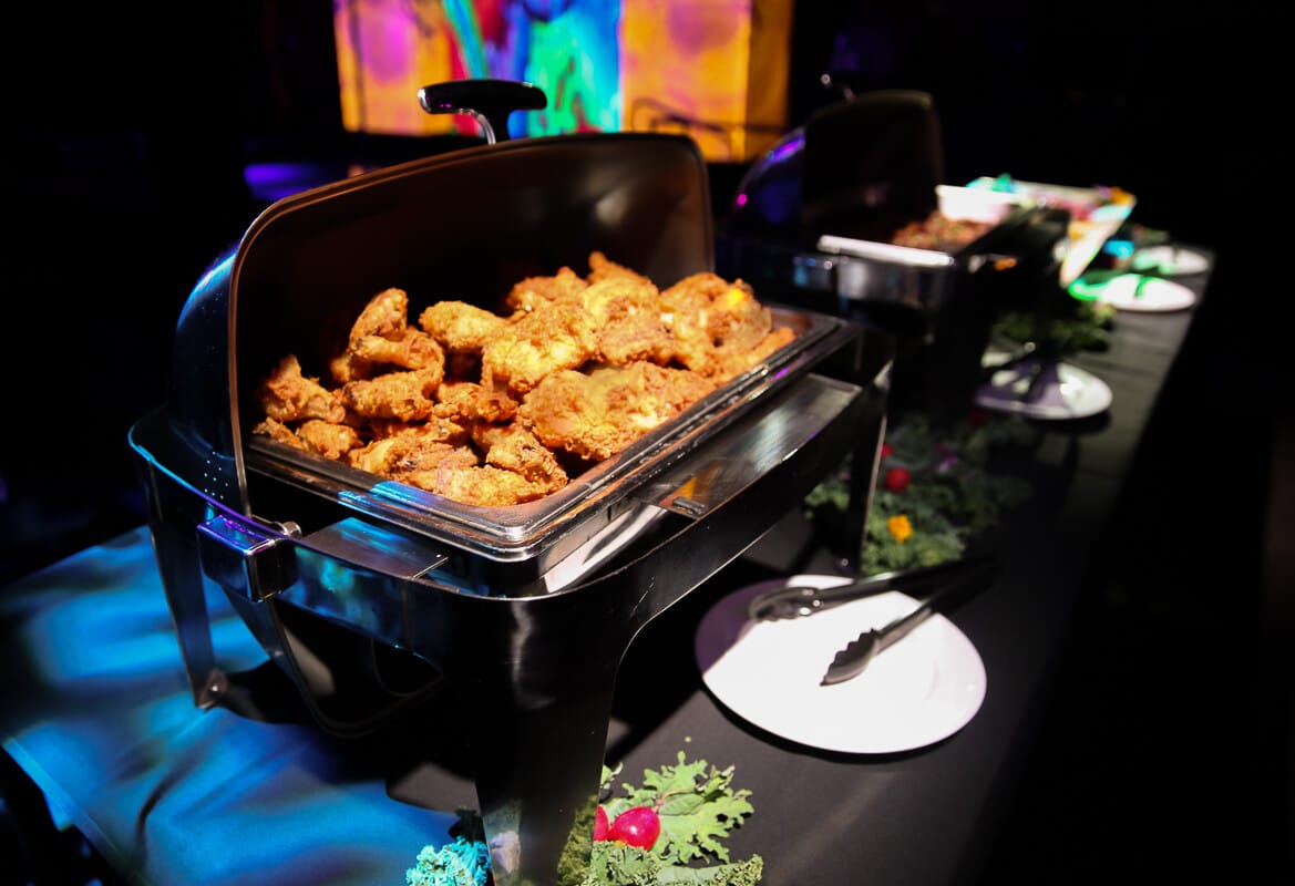 A buffet-style tray filled with crispy fried chicken is placed on a table. There's a serving tong beside a white plate, and colorful lights illuminate the scene.