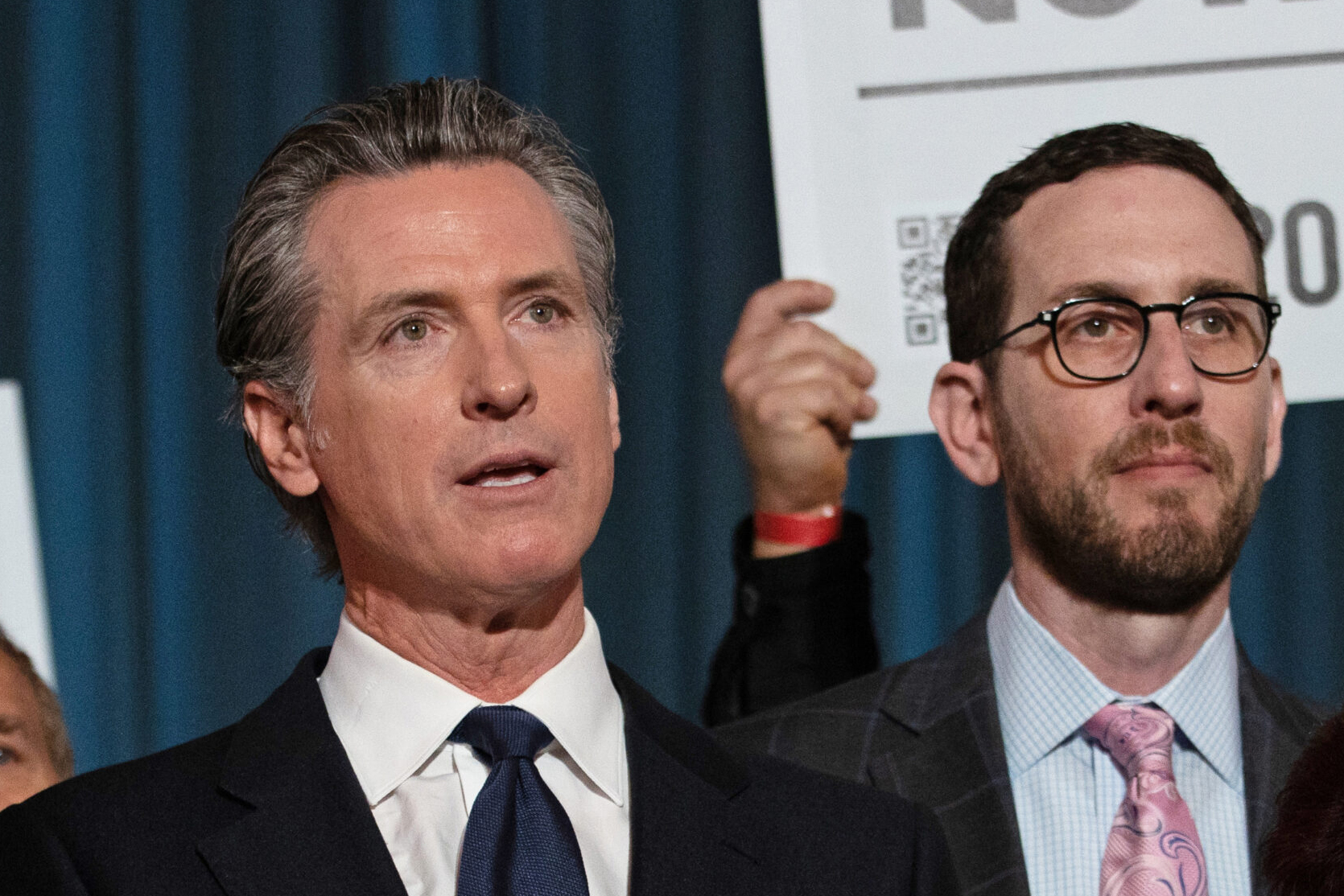 Two men in suits are standing in front of a blue curtain. One holds a sign partially visible in the background. They appear to be engaged in a serious discussion.