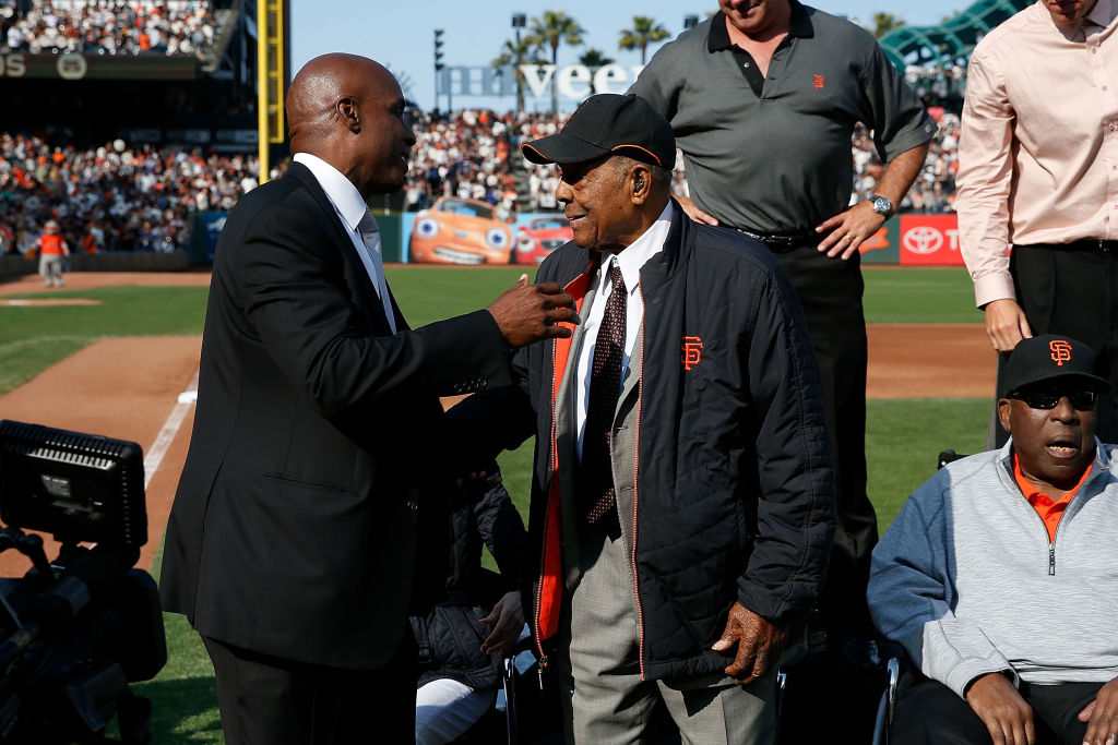 Two men stand on a baseball field with a crowd in the background. They appear to be greeting each other warmly. Two other men, one seated, are nearby.