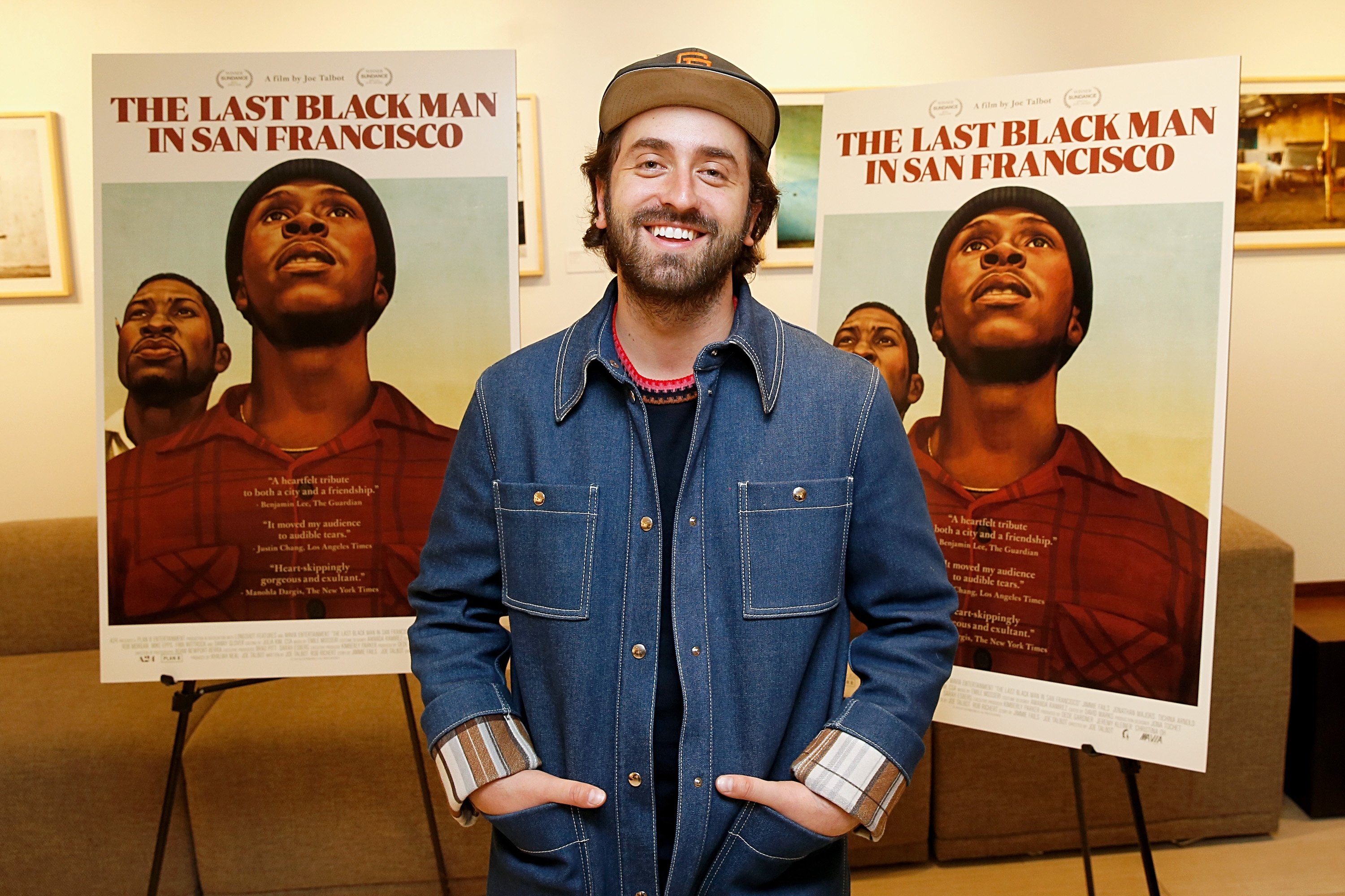 A person in a denim jacket and cap smiles in front of two posters for "The Last Black Man in San Francisco," featuring two men looking upwards.