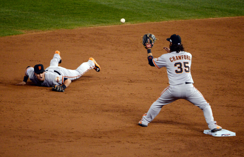 Two baseball players in grey uniforms are on a dirt field. One dives flat on the ground, while the other stands ready to catch a ball near a base.