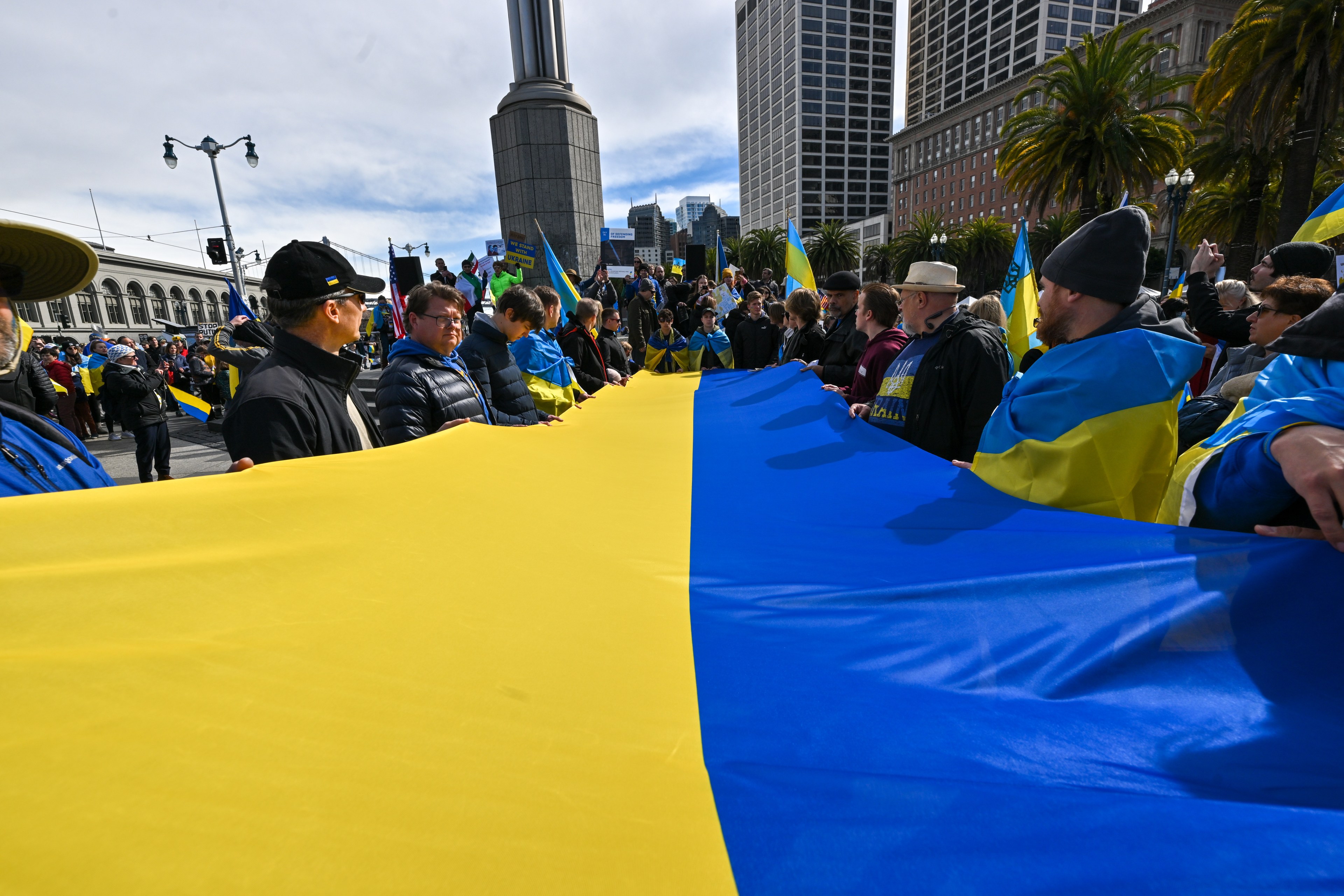 A group of people is holding a large Ukrainian flag in a public area, surrounded by more individuals with smaller flags. Buildings and palm trees are in the background.