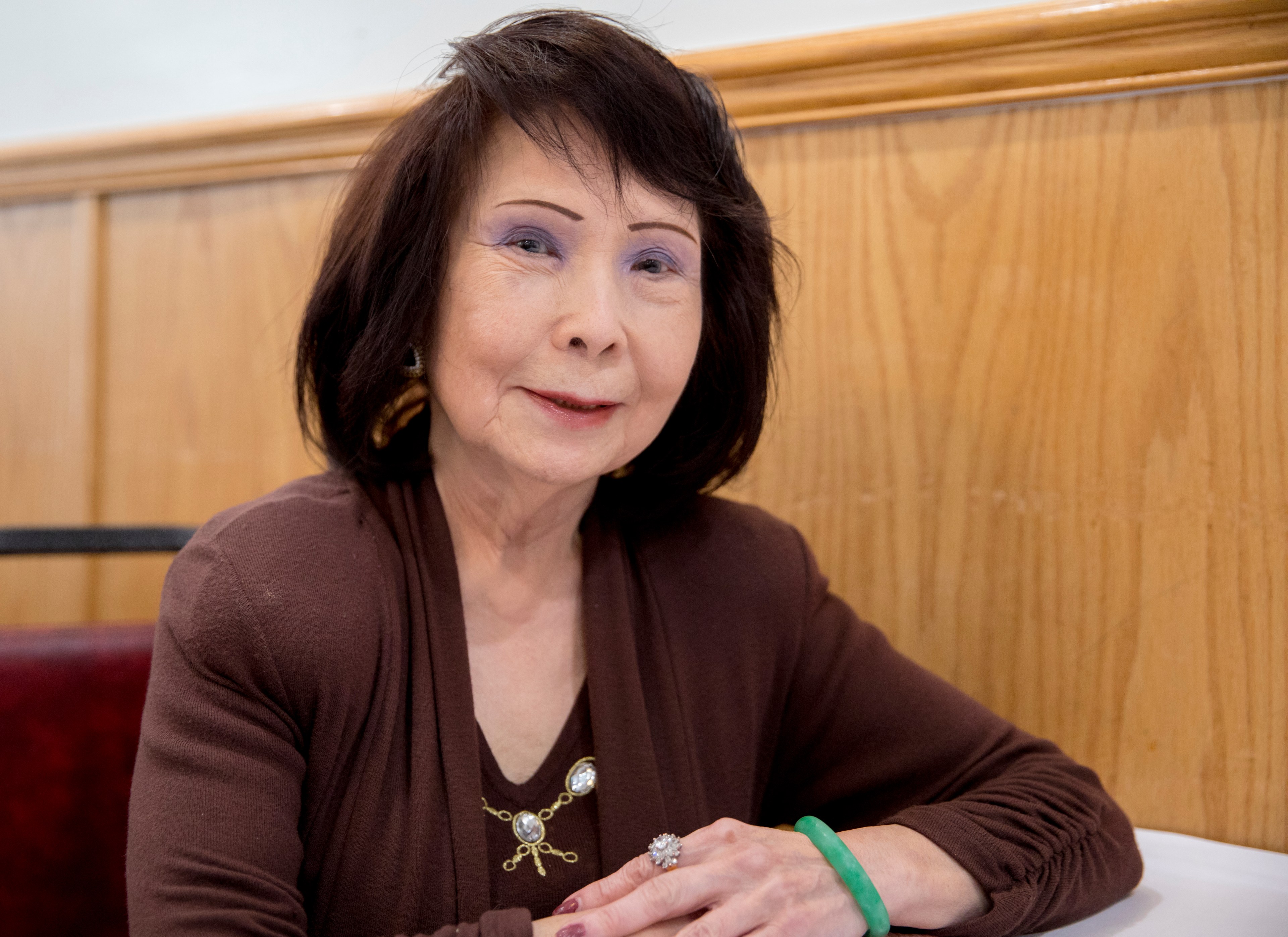 A woman with dark hair sits smiling, wearing a brown top with a decorative brooch. She has a green bracelet and a ring, and there's wood paneling behind her.