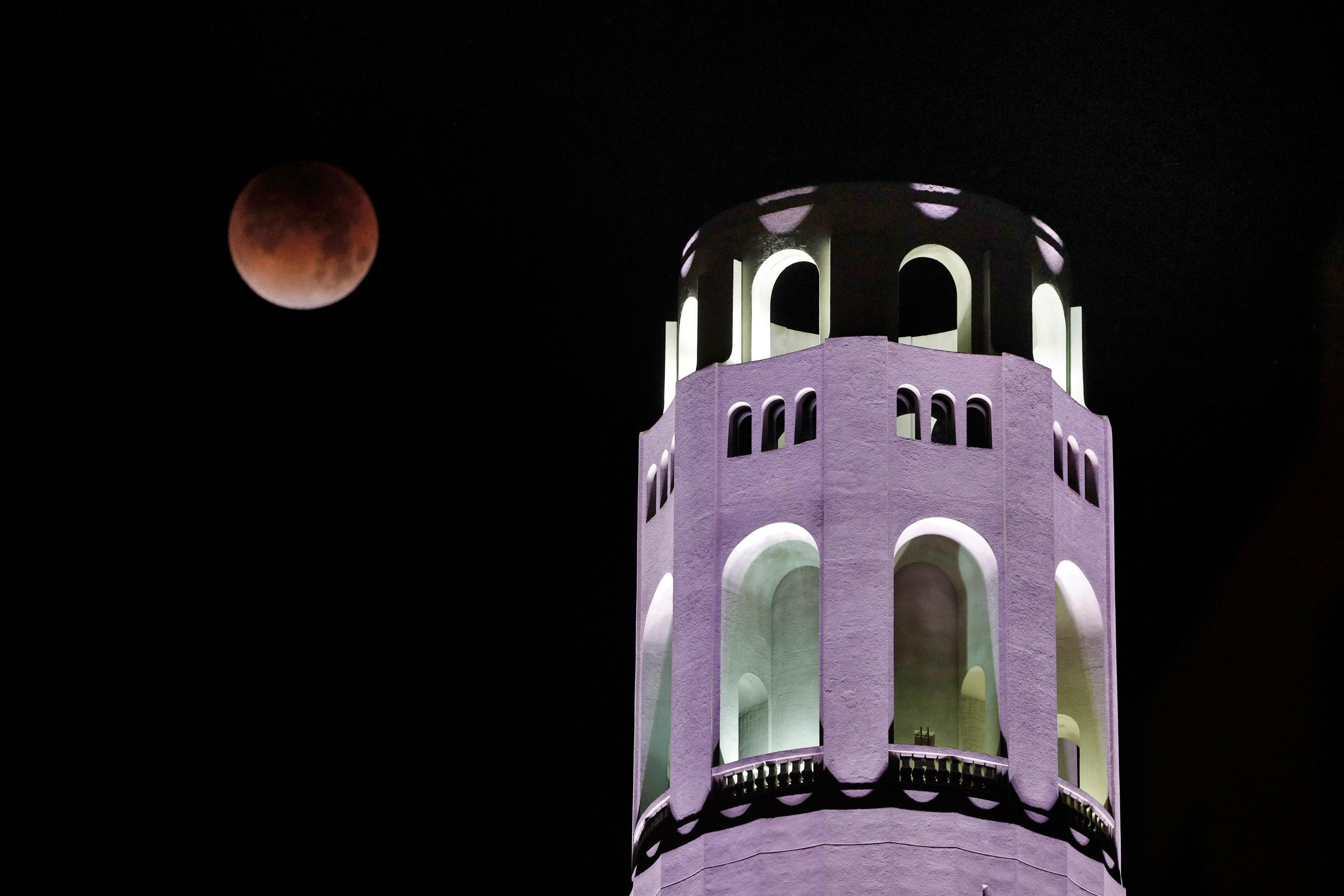 A blood moon glows in the dark sky near a well-lit, white, cylindrical tower with arched windows.