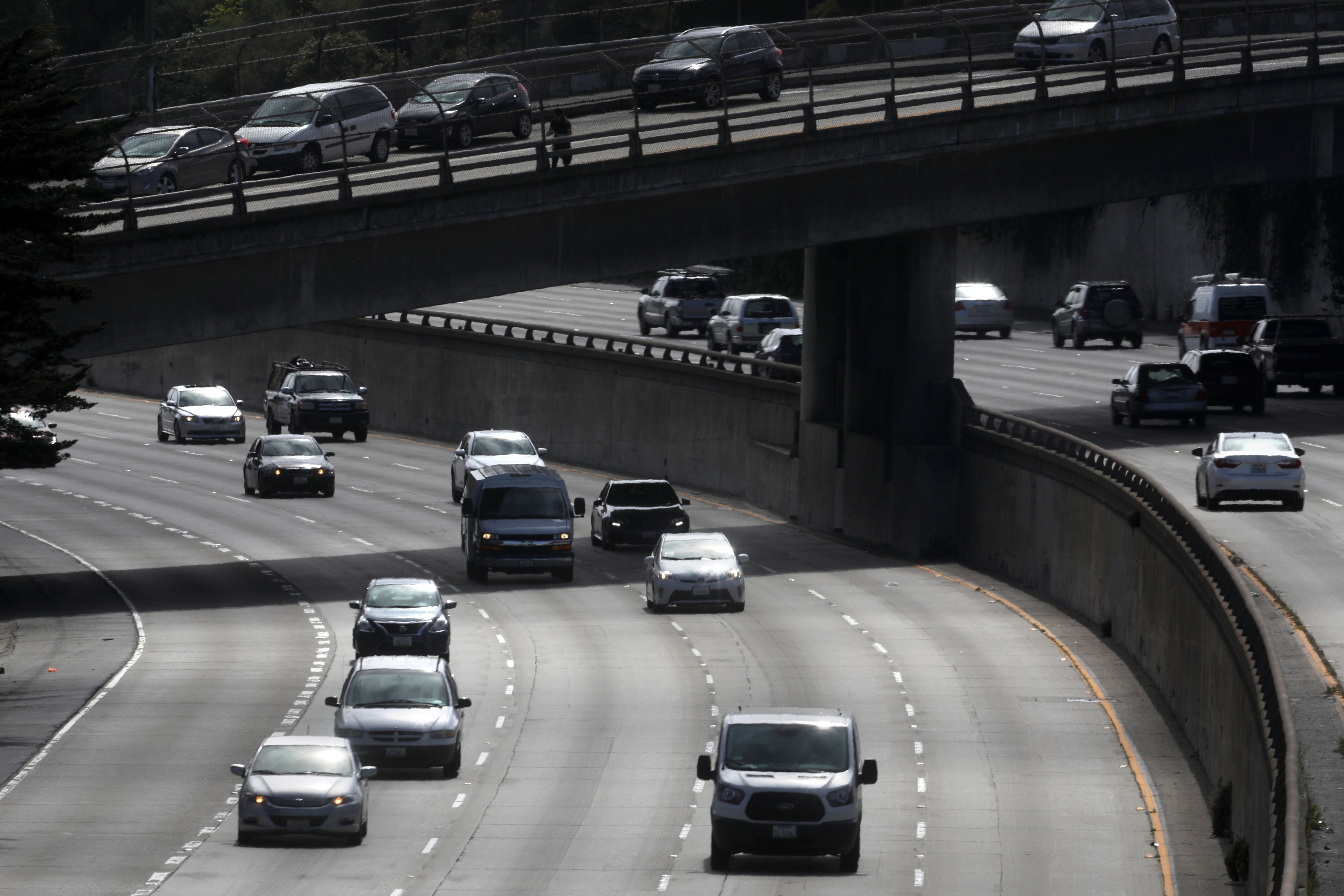 The image shows a highway with multiple lanes, featuring several cars moving along. There's an overpass above, carrying more vehicles in the opposite direction.