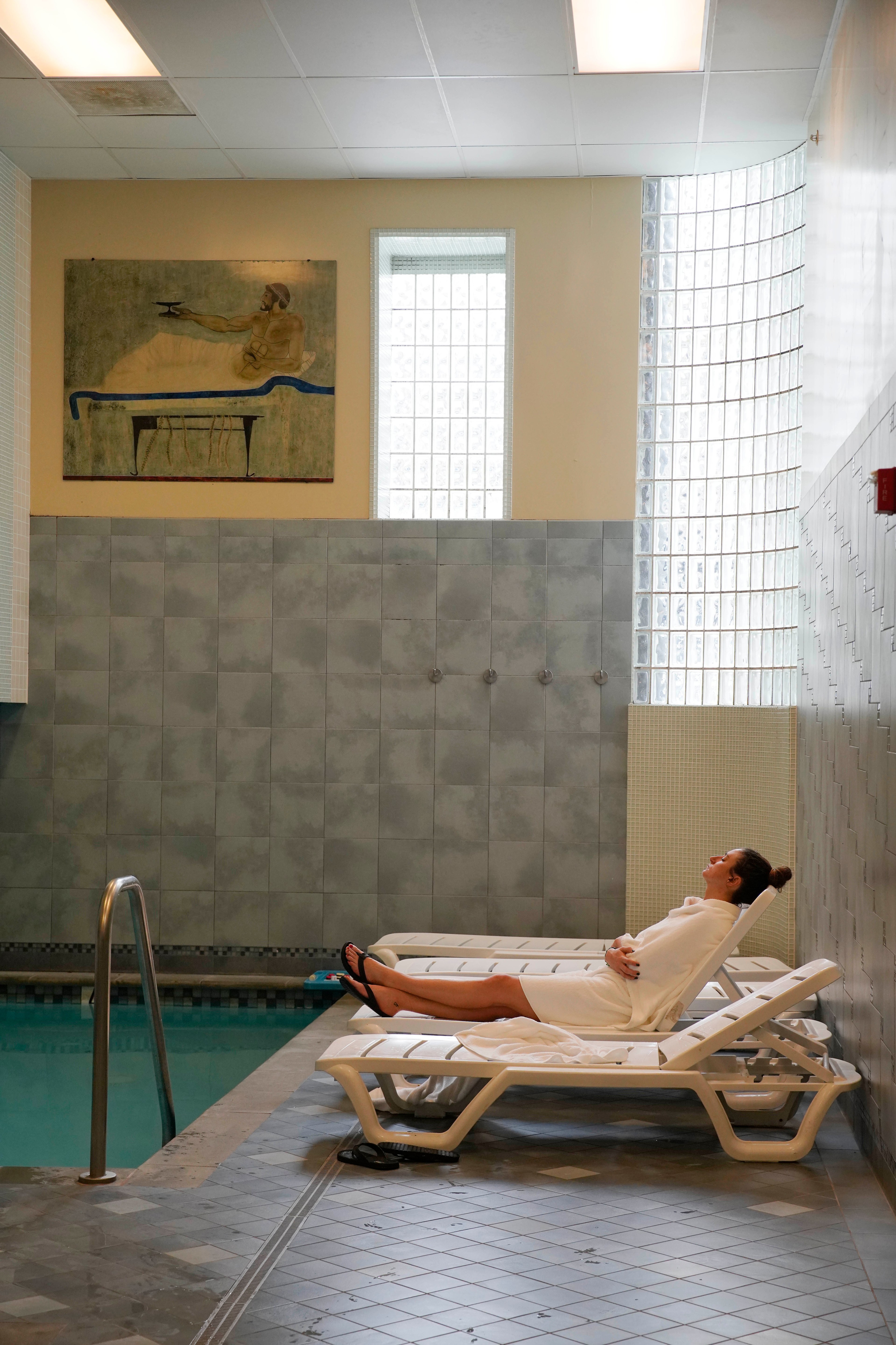 A person in a white robe relaxes on a lounge chair beside an indoor pool. The room has tiled walls, a high ceiling, and a painting on the wall.