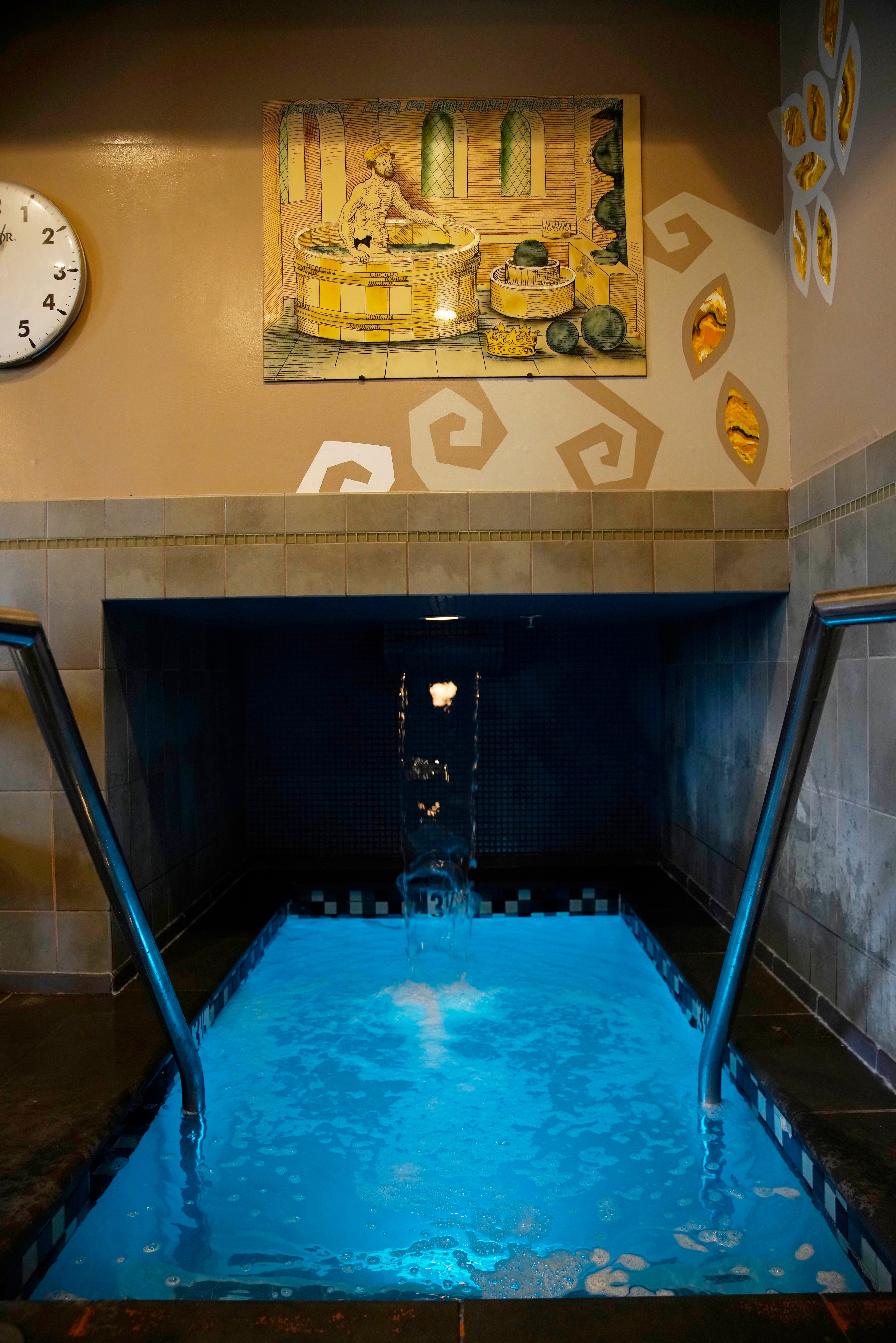 A small indoor pool with clear blue water features two handrails and a waterfall. Above, a painting depicts a person in a vintage bath setting. A clock shows 4:05.