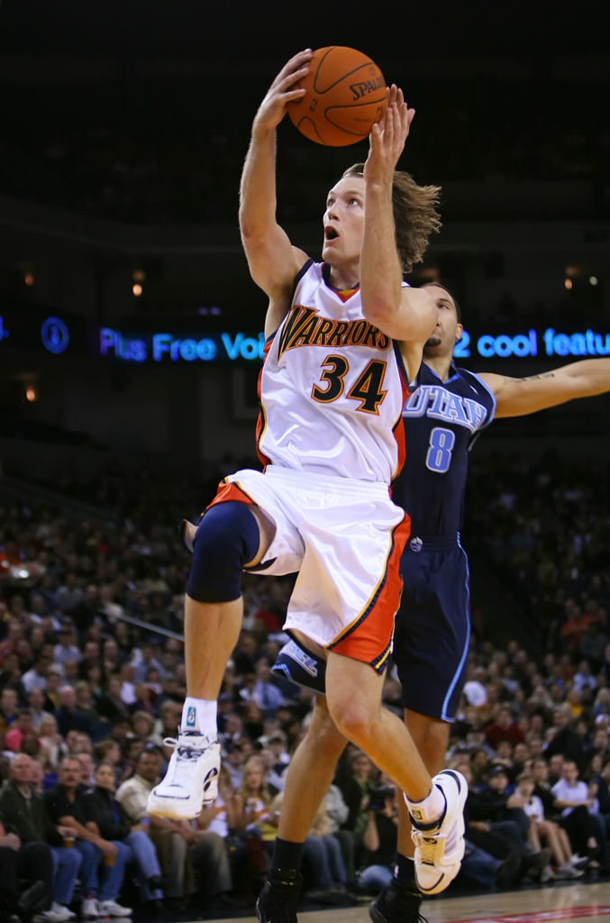 A basketball player in a white Warriors jersey is mid-air, holding the ball above him, with another player in a dark Utah jersey attempting to defend.