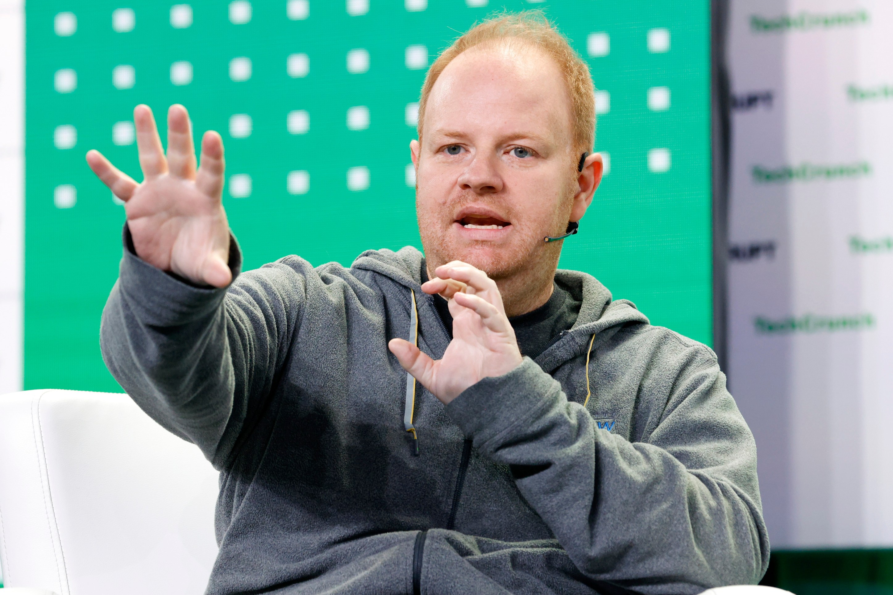 A person in a gray hoodie is speaking on stage with a microphone. The background is green with white squares and partially visible text.