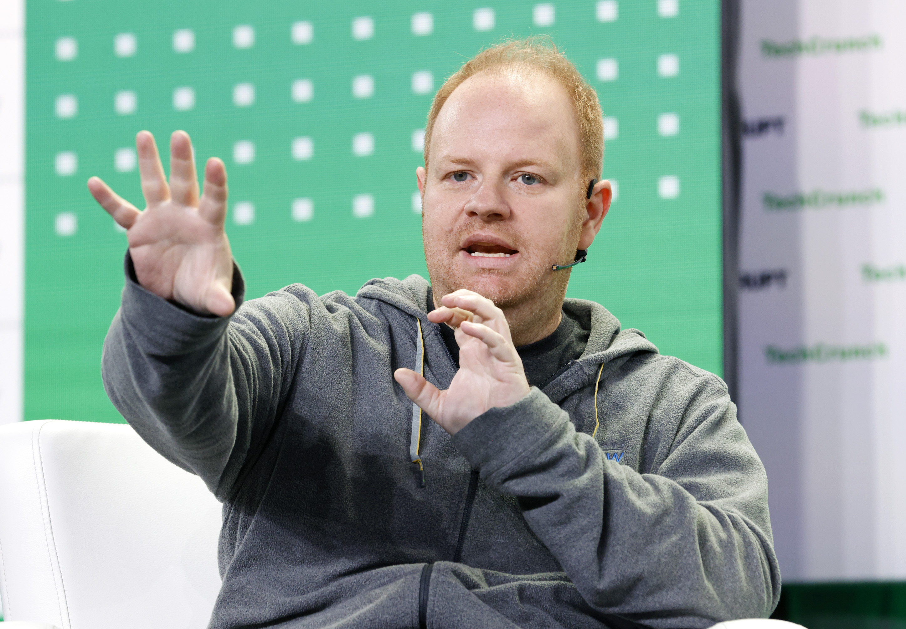 A person in a gray hoodie is speaking on stage with a microphone. The background is green with white squares and partially visible text.