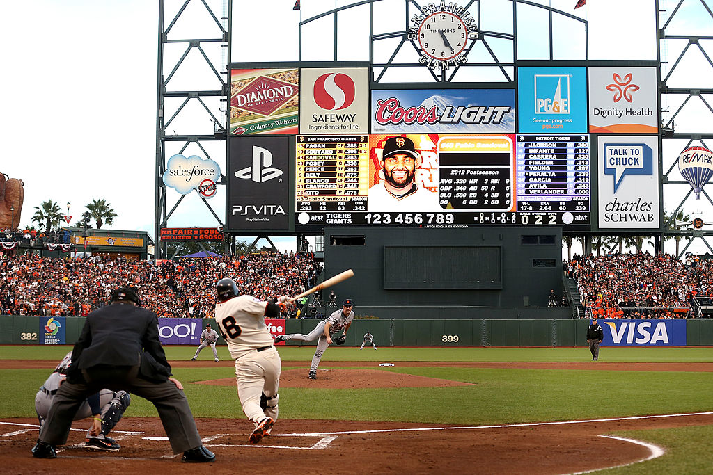 The image shows a baseball player swinging at a pitch in a crowded stadium. A large scoreboard displays player stats, surrounded by various advertisements.