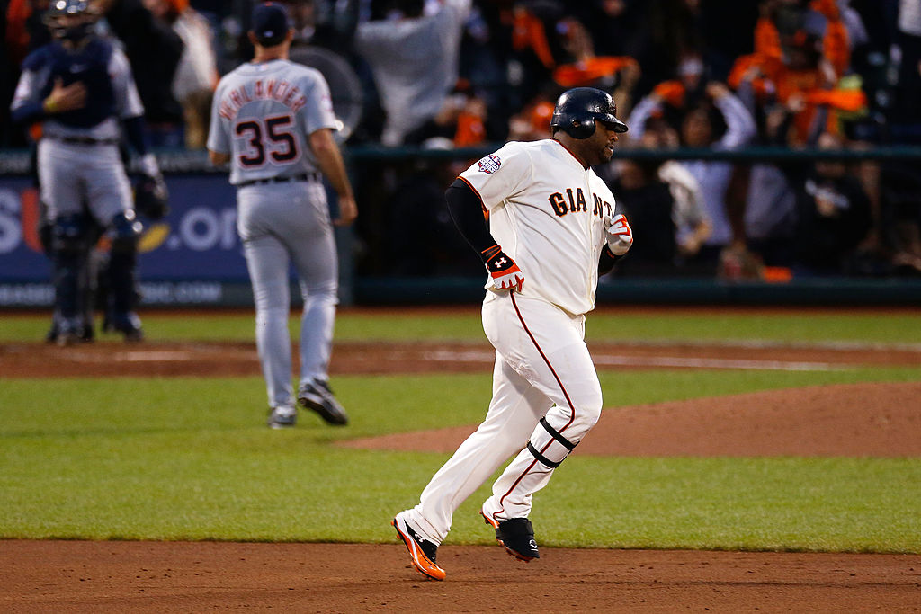 A baseball player in a Giants uniform is running, likely after a hit. Two opponents in gray uniforms stand in the background, with a crowd watching intently.