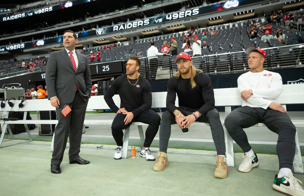 Four men are by a football field; one is in a suit, and three in casual sportswear and hats, sitting on a bench. The stadium is partially filled.