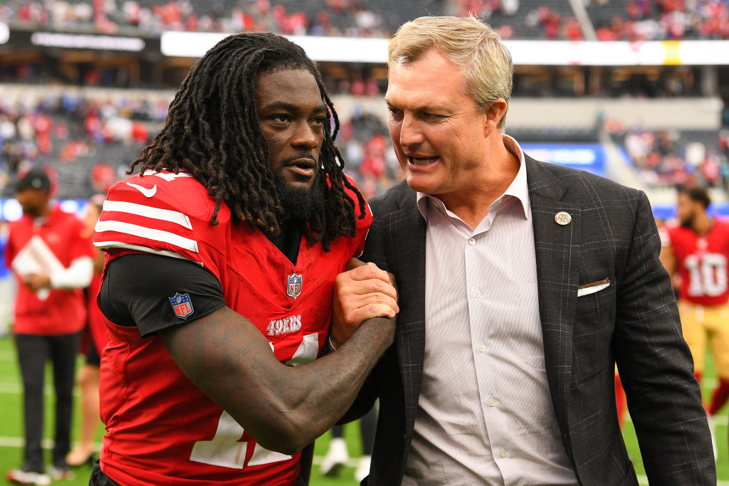 A football player in a red jersey shakes hands with a man in a suit on a field, with blurred spectators in the background.